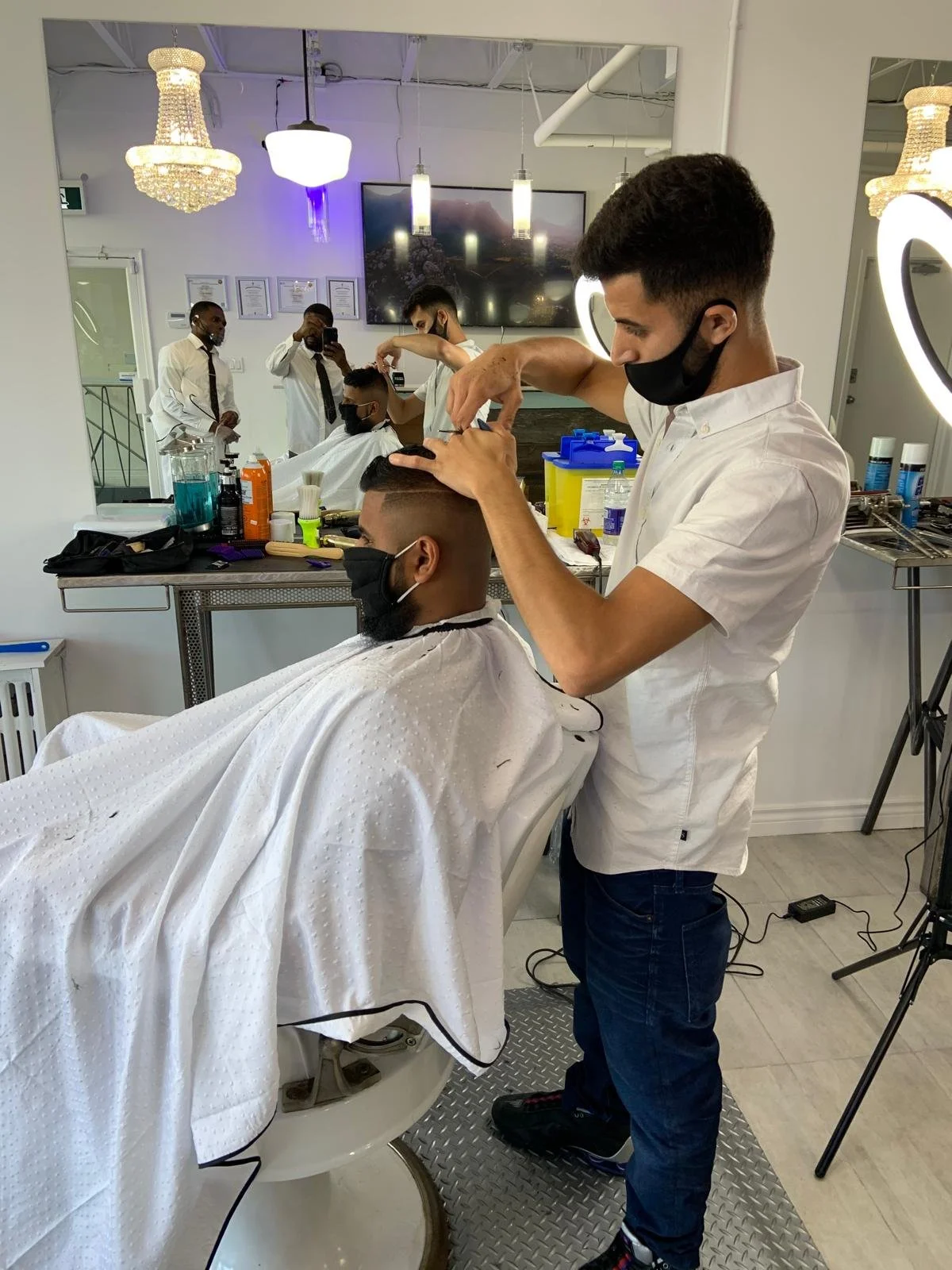 A barber is cutting a customer's hair in a salon with multiple barber stations. The barber is wearing a white shirt and black mask, and the customer is seated with a cape, also wearing a black mask. There are other barbers and customers in the backgr