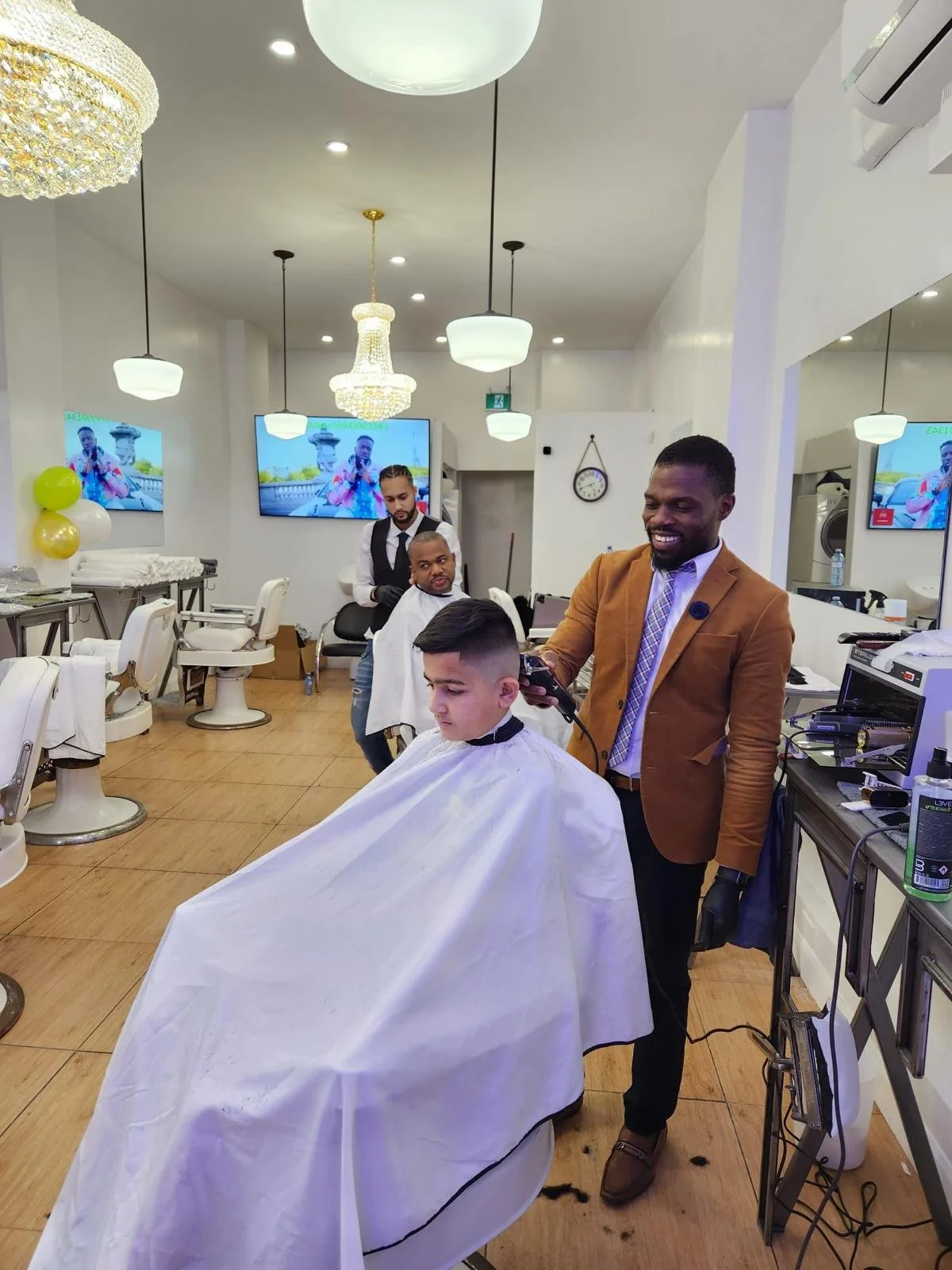 A man in a brown blazer is giving a young man a haircut at a barbershop, with other clients and barbers in the background, decorated with chandeliers and wall-mounted TVs.