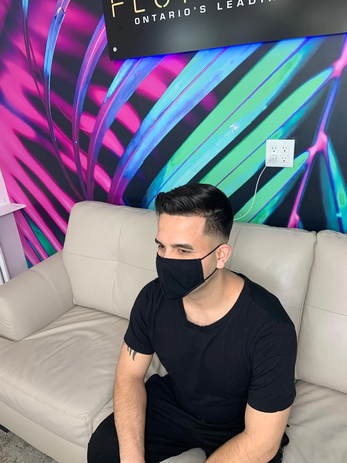 A man wearing a black face mask and black t-shirt sitting on a beige sofa in front of a colorful mural with neon-like pink, blue, and green lines and patterns.