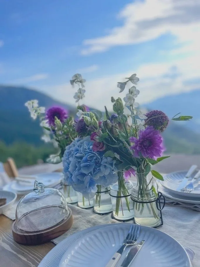 Outdoor dining table set with white plates, cutlery, and glass jars filled with purple, blue, and white flowers, against a mountain and sky backdrop.