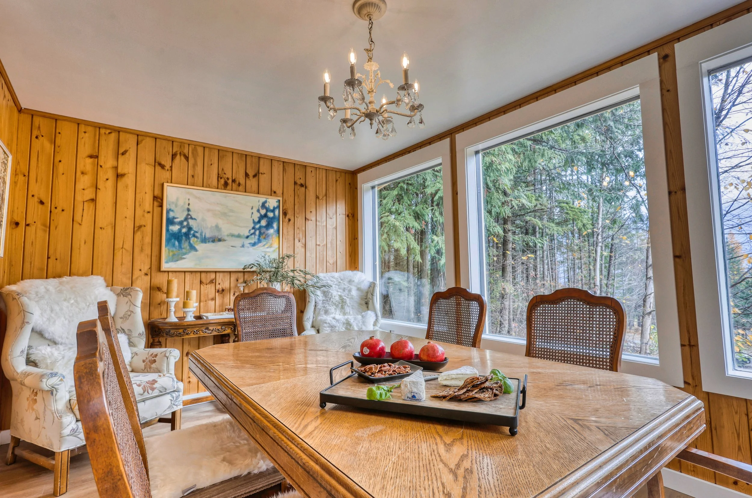Cozy dining room with wooden walls, a chandelier, a large window showing a forest view, and a table set with pomegranates and cheese.