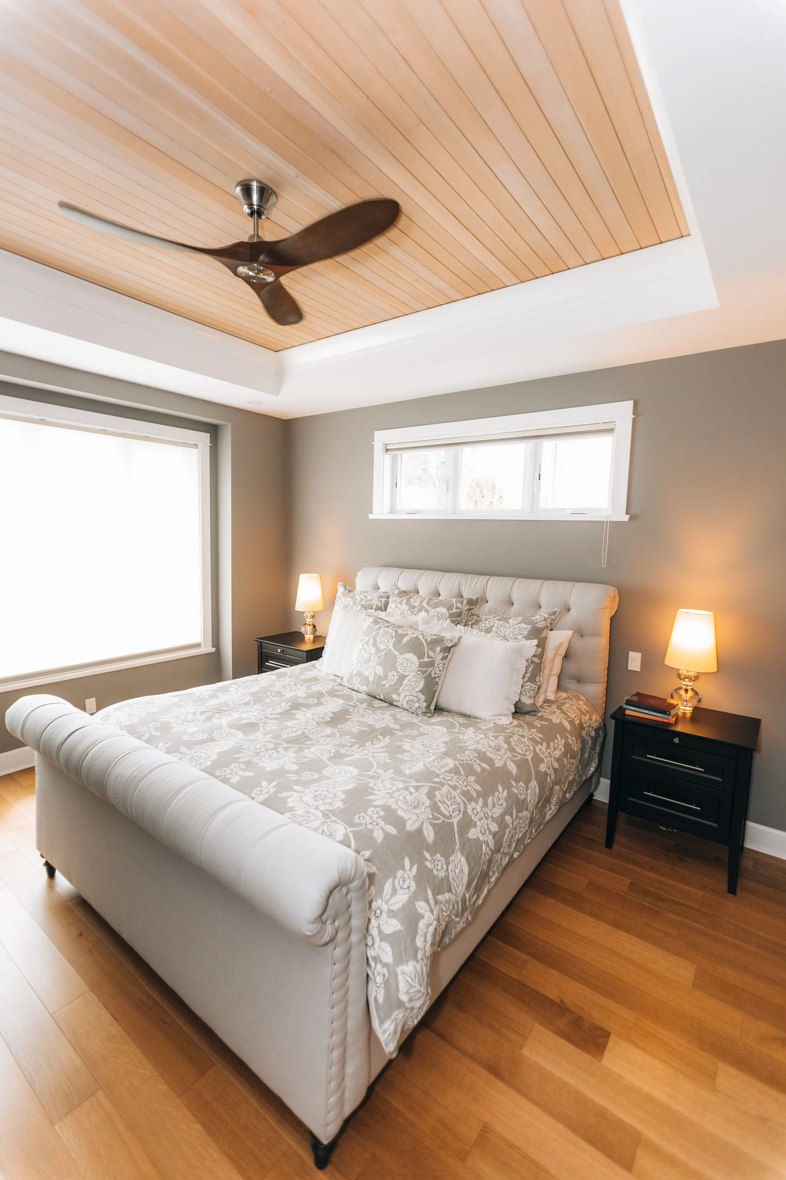 Modern bedroom with a bed featuring floral bedding, two bedside tables with lamps, wooden ceiling with a ceiling fan, and natural light from windows.