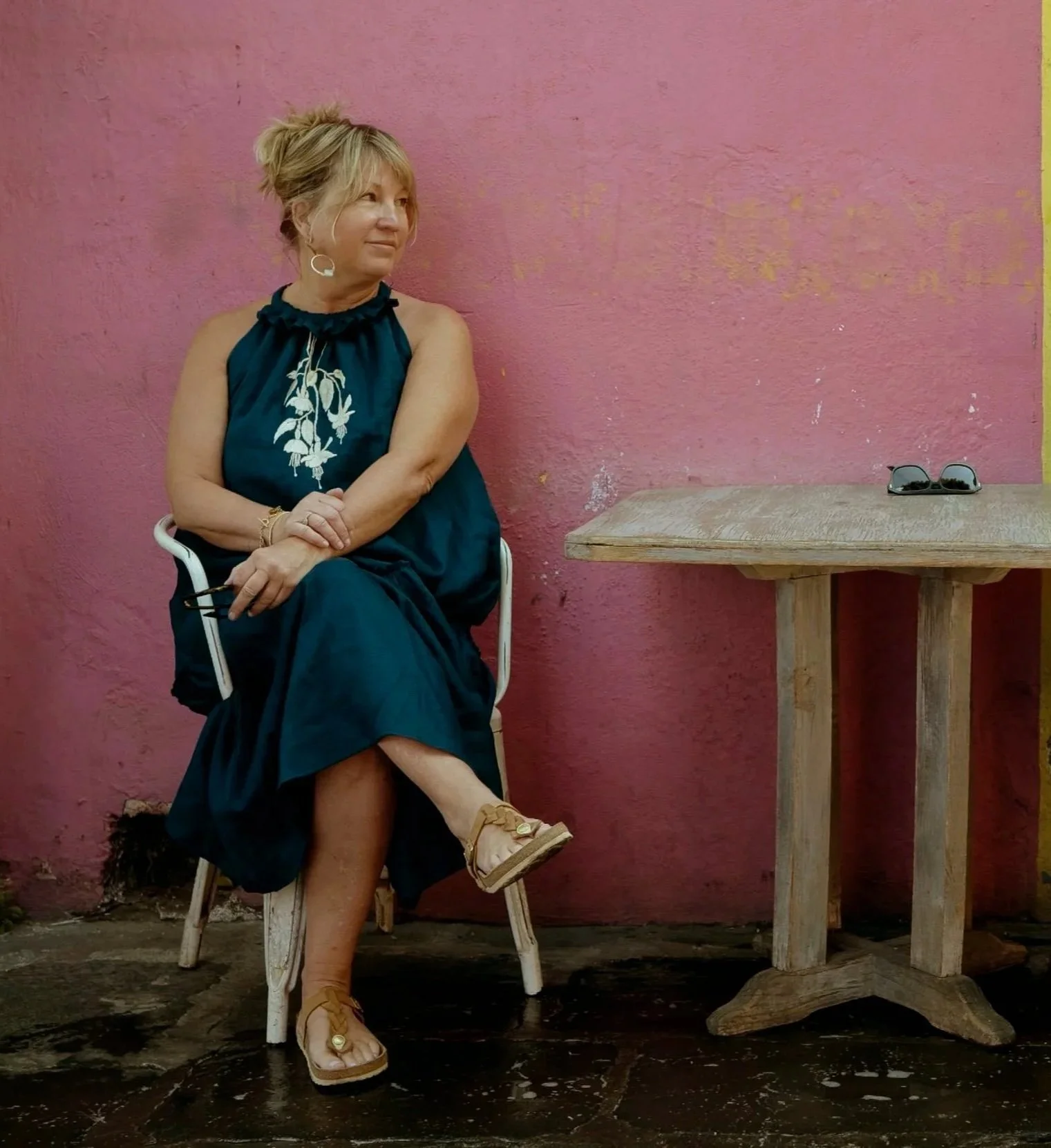 A woman in a dark blue dress with white embroidery sitting on a white chair against a pink wall, with sunglasses on a wooden table beside her.