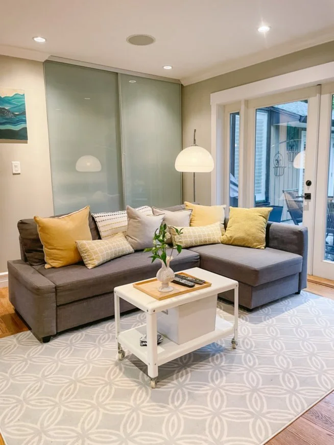 Cozy living room with gray sectional sofa, yellow and beige pillows, small white coffee table, decorative vase, remote controls, modern floor lamp, sliding glass door, patterned rug, and artwork on the wall.