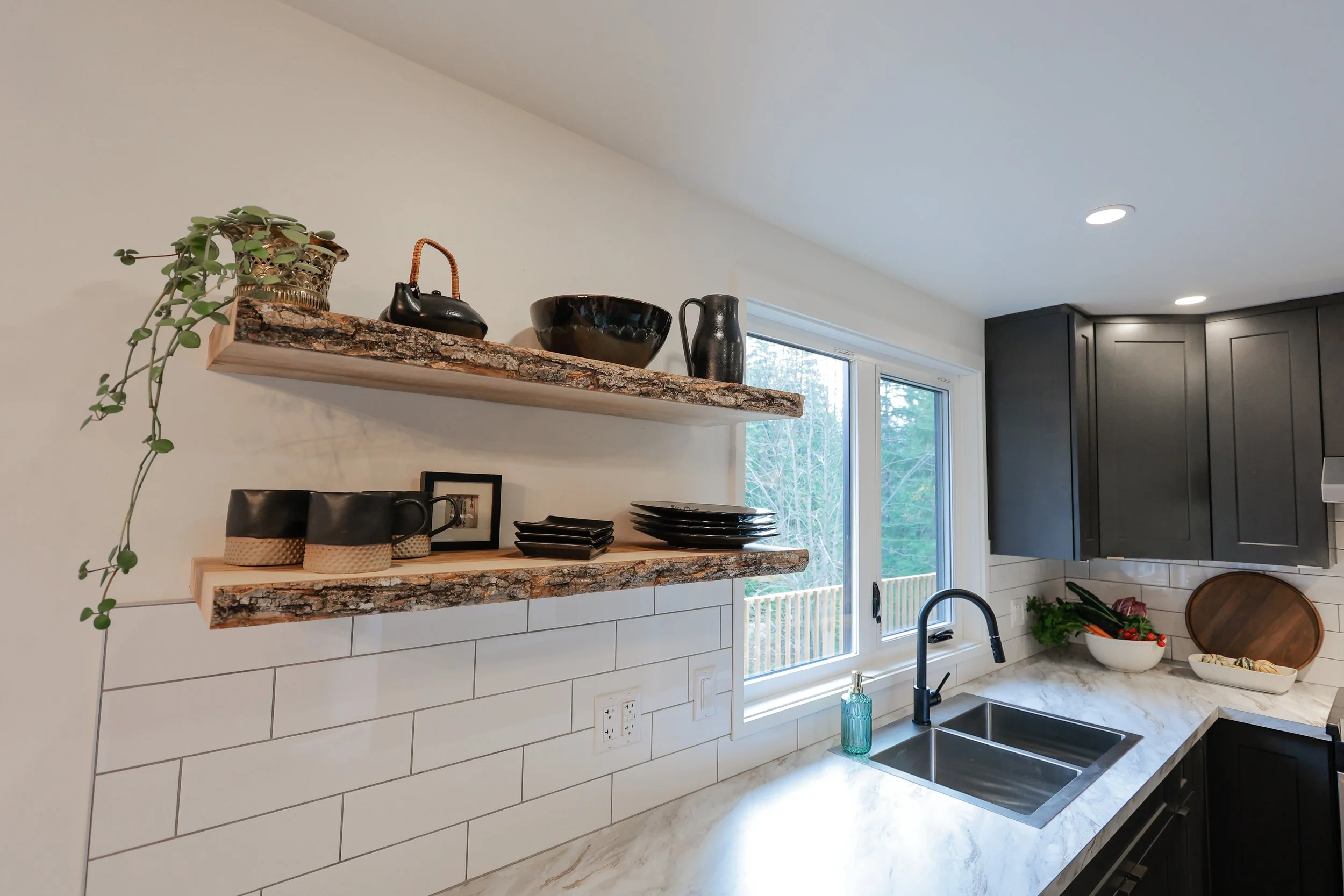Modern kitchen with white subway tile backsplash, live-edge wood shelves holding black ceramic dishes and a plant, black faucet, and gray cabinets. Natural light from window and countertop with sink and decorative items.