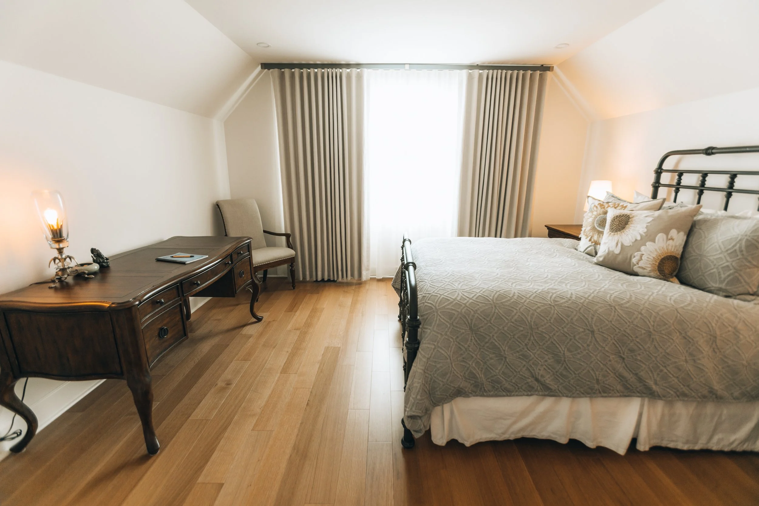 Cozy bedroom with a large bed, floral pillows, wooden desk, chair, and curtain-covered window.
