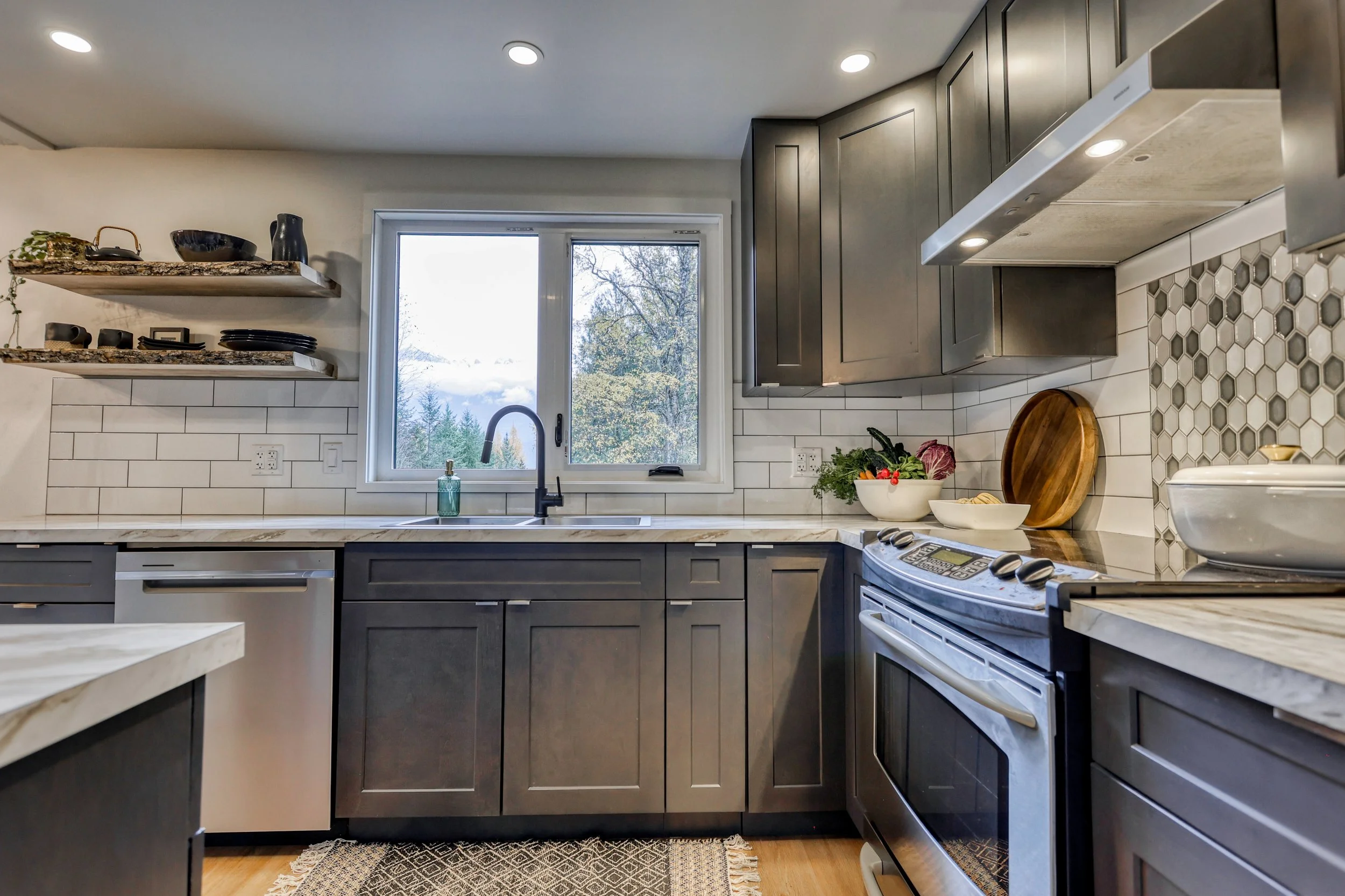 Modern kitchen interior with gray cabinets, white tile backsplash, and stainless steel appliances. Open shelves hold black dishes, and a window above the sink offers a view of trees. Countertops feature a cutting board, bowls of produce, and a pot on