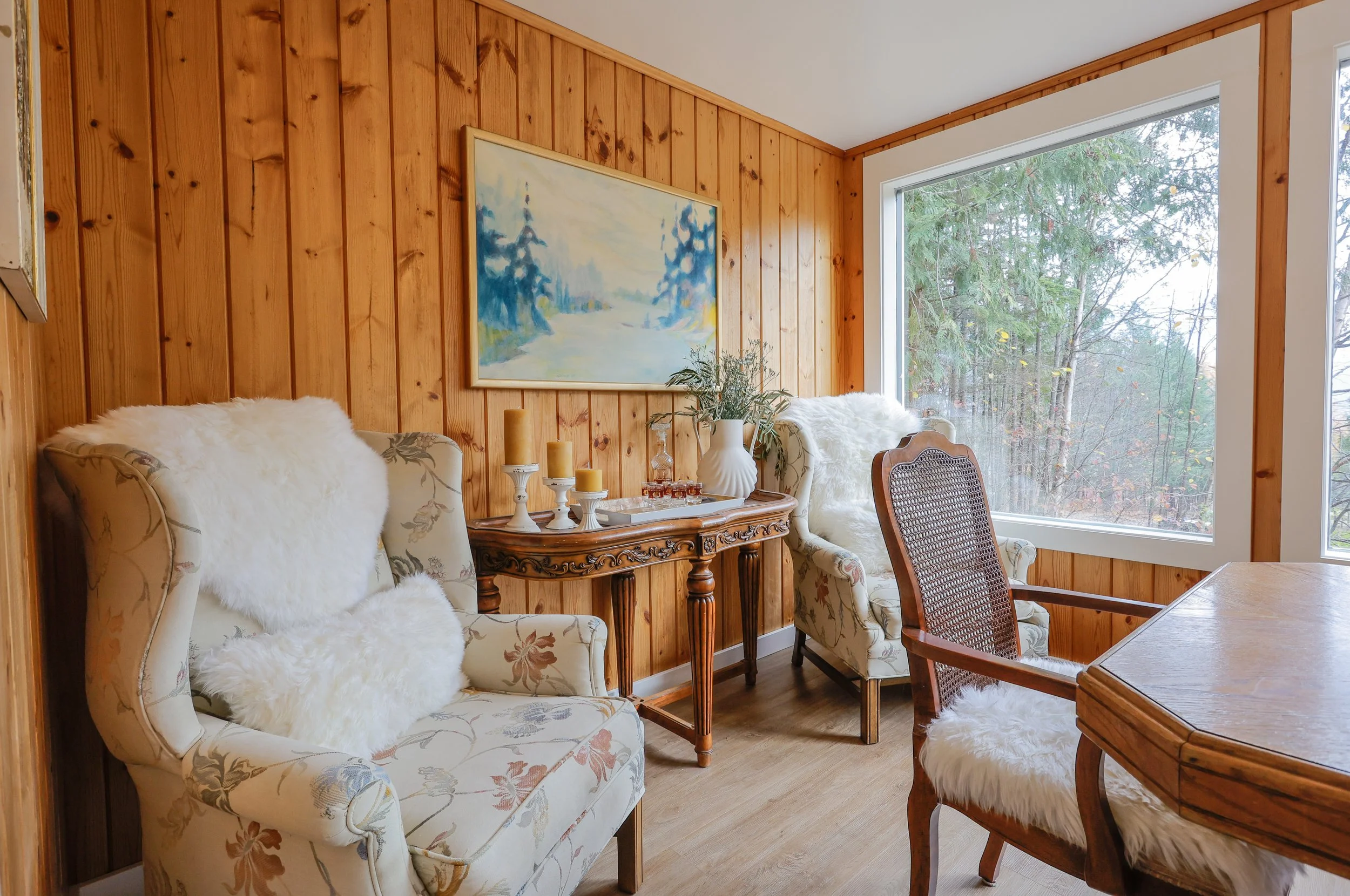Cozy room with wooden paneling, two floral armchairs with white fur throws, a wooden table with candles and decor, a landscape painting on the wall, and large window with view of trees.
