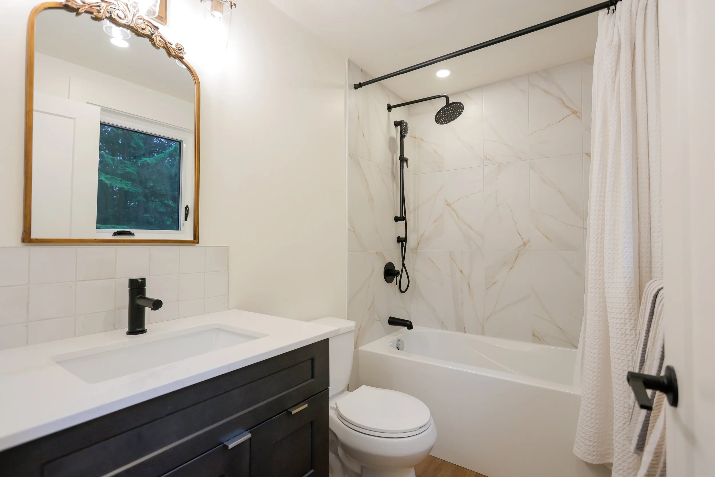 Modern bathroom with bathtub, shower, black fixtures, marble tiles, vanity with sink, mirror, and towel rack.