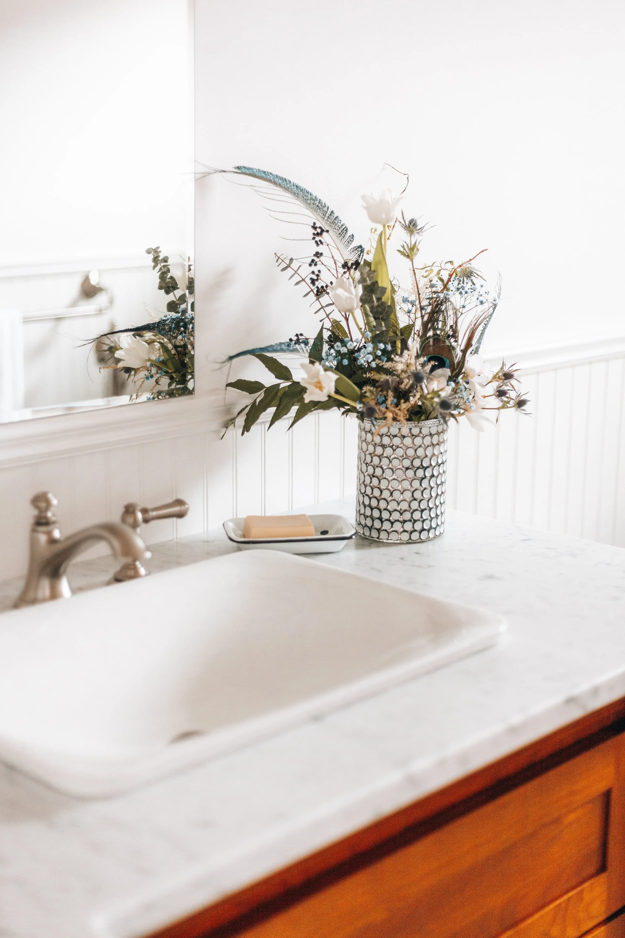 Bathroom sink area with a white marble countertop, chrome faucet, decorative floral arrangement in a textured vase, bar of soap on a dish, and a wall mirror.