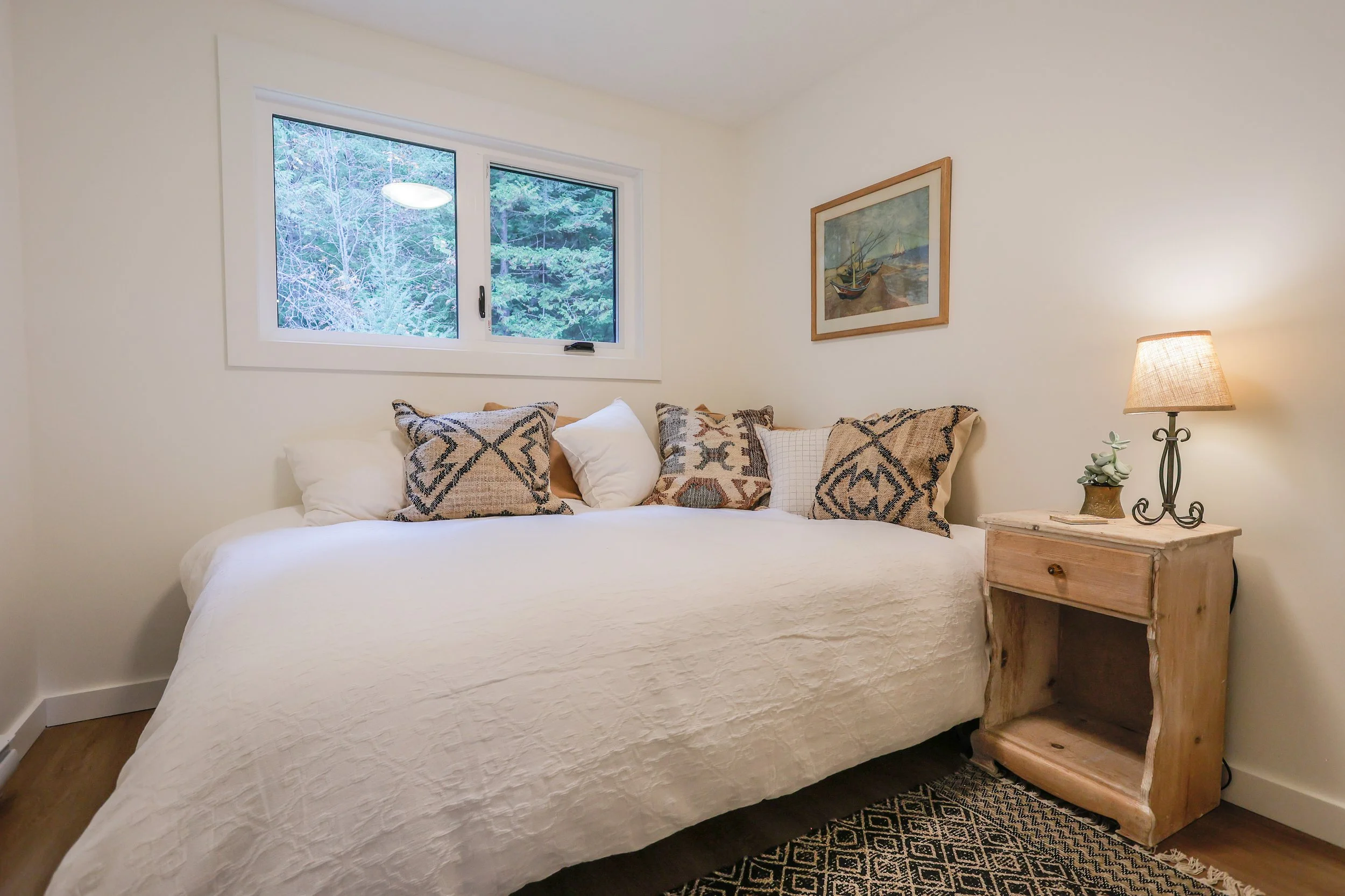 Small cozy bedroom with a large bed covered in white linens and patterned pillows. There's a wooden nightstand with a lamp and a small plant. A window overlooking greenery and a framed picture on the wall complete the decor.