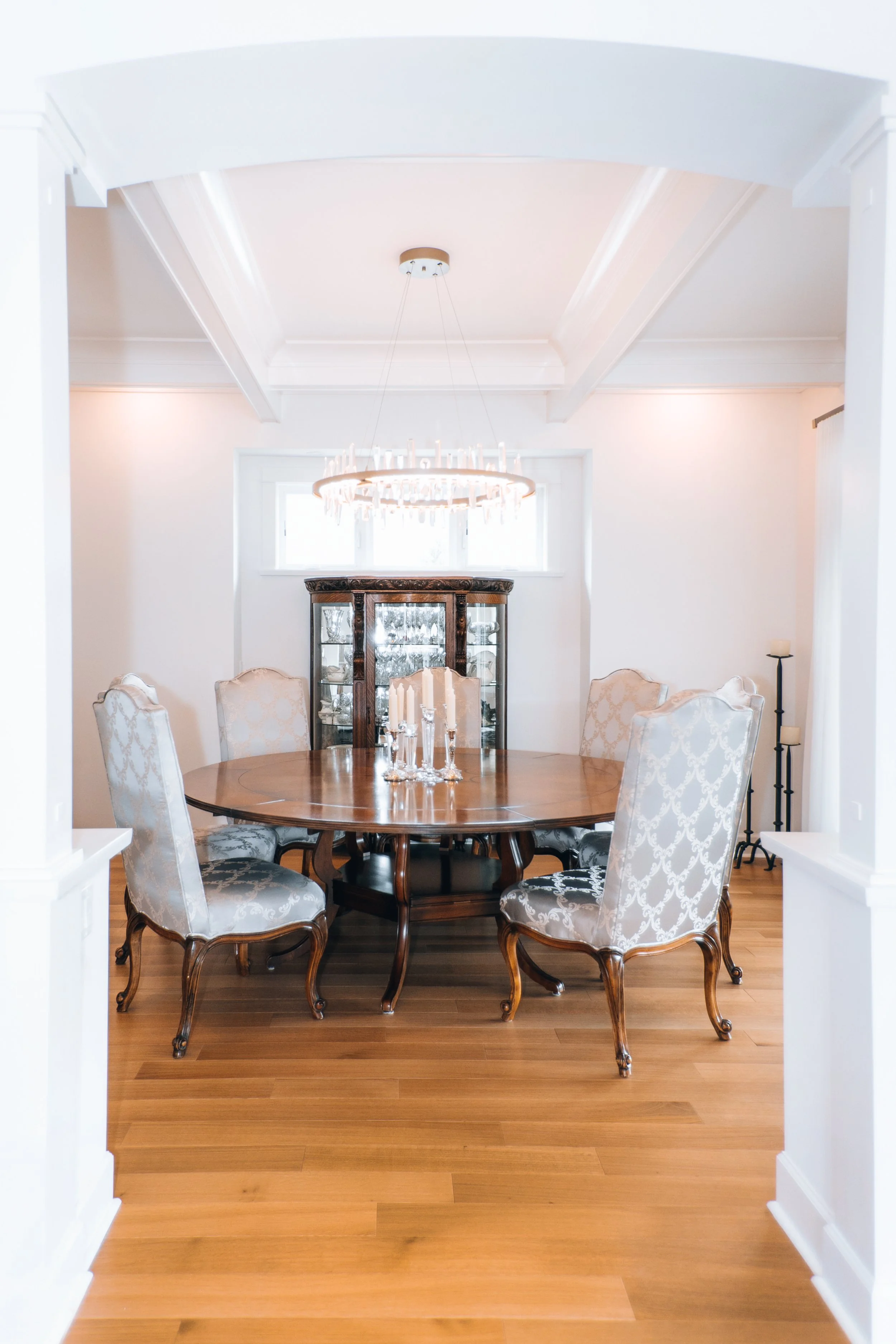 Elegant dining room with a round wooden table, upholstered chairs, chandelier, and glass cabinet.