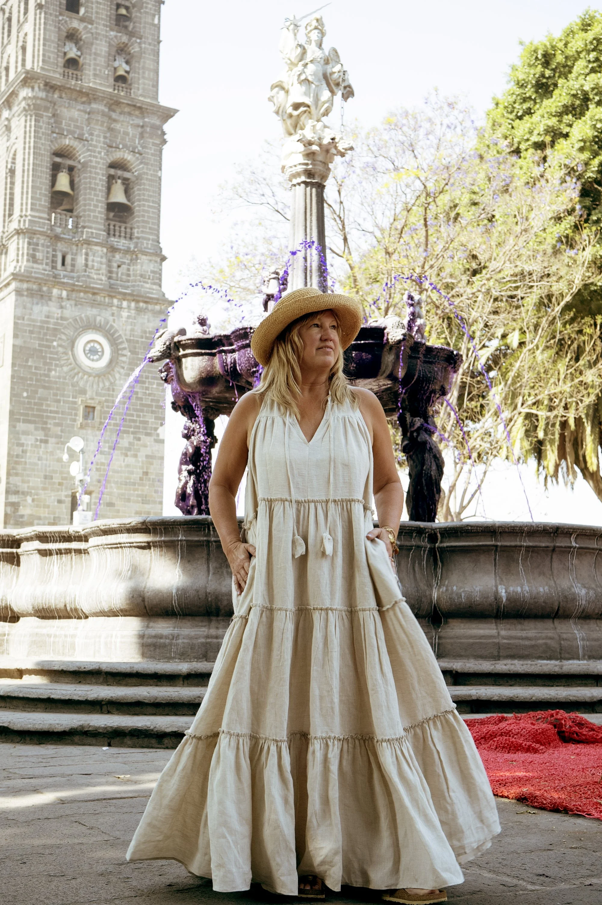 A woman in a long beige dress and a wide-brimmed straw hat stands in front of a fountain with purple water in an outdoor setting, with a historic building and trees in the background.