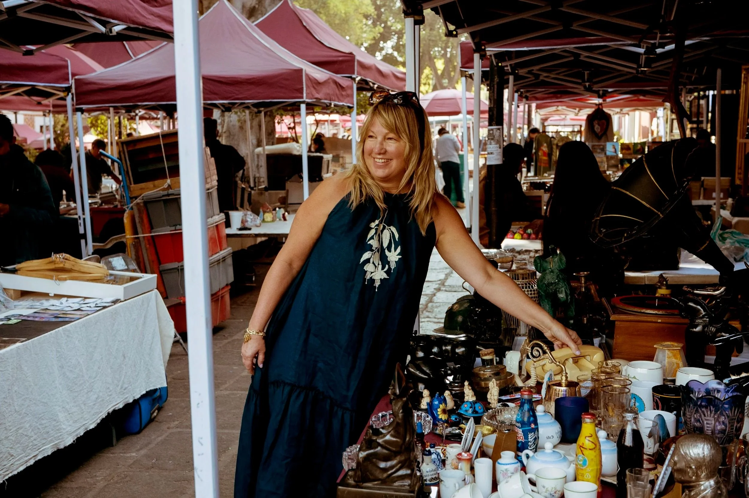 A woman shopping at an outdoor flea market with various items like ceramics, glassware, and decorative objects on display under maroon canopies.