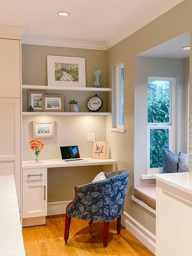 Home office nook with built-in desk, laptop, decorative vases, clock, and artwork on shelves, blue patterned chair, window with cushioned seat, and flowers in a vase.