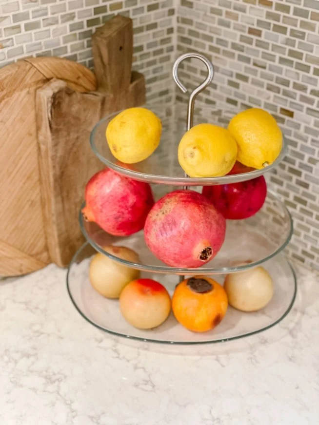 Three-tiered glass fruit stand with lemons, pomegranates, and persimmons on a kitchen countertop with a wooden cutting board and mosaic tile backsplash.