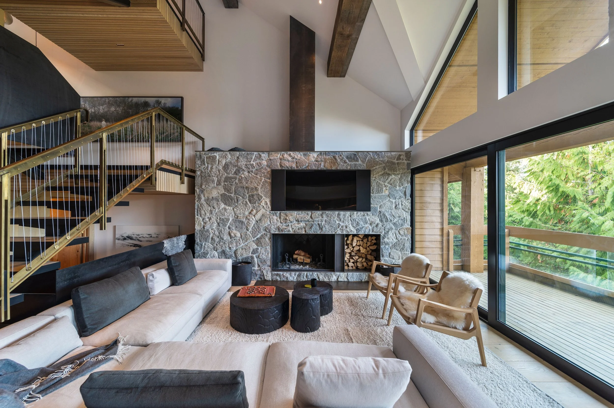 Modern living room with a stone fireplace, large windows, and a cozy seating area. Features include a sectional sofa, wooden chairs with plush cushions, a staircase, and a stack of firewood. The room is bright with natural light from the windows, off