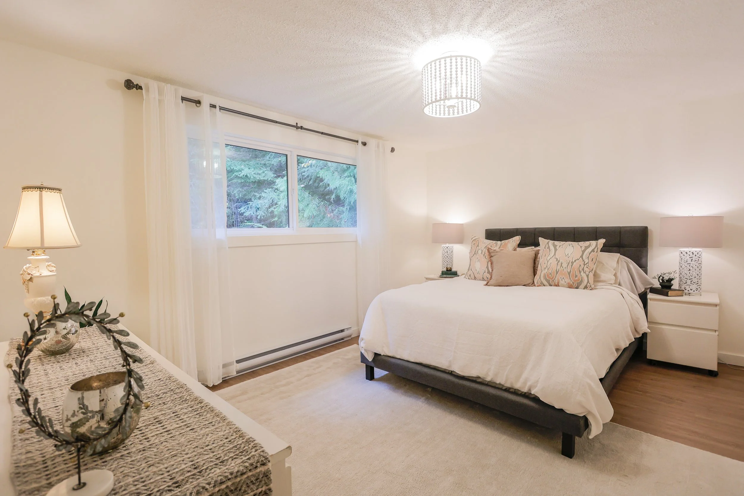 Modern bedroom with a large bed, white bedding, decorative pillows, two bedside tables with lamps, a window with sheer curtains, and a ceiling light.