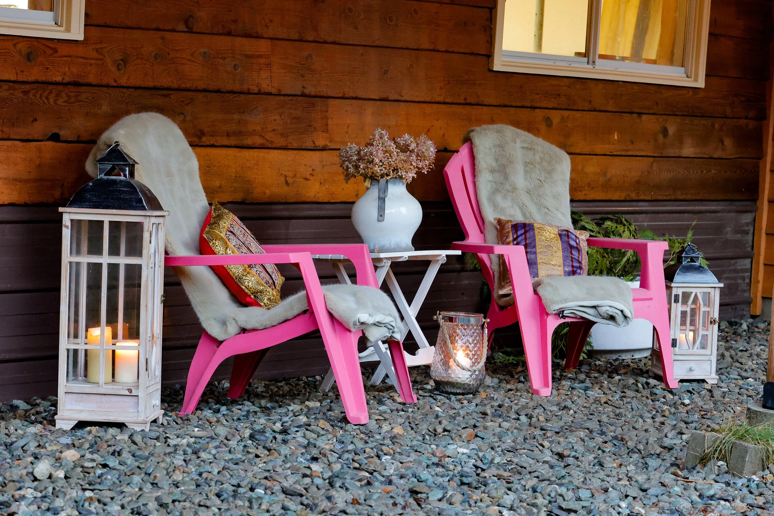 Two pink outdoor chairs with fur throws and decorative pillows, flanking a small white table with a floral arrangement in a white vase. Large lanterns with candles are on either side, set on a gravel surface next to a wooden wall.