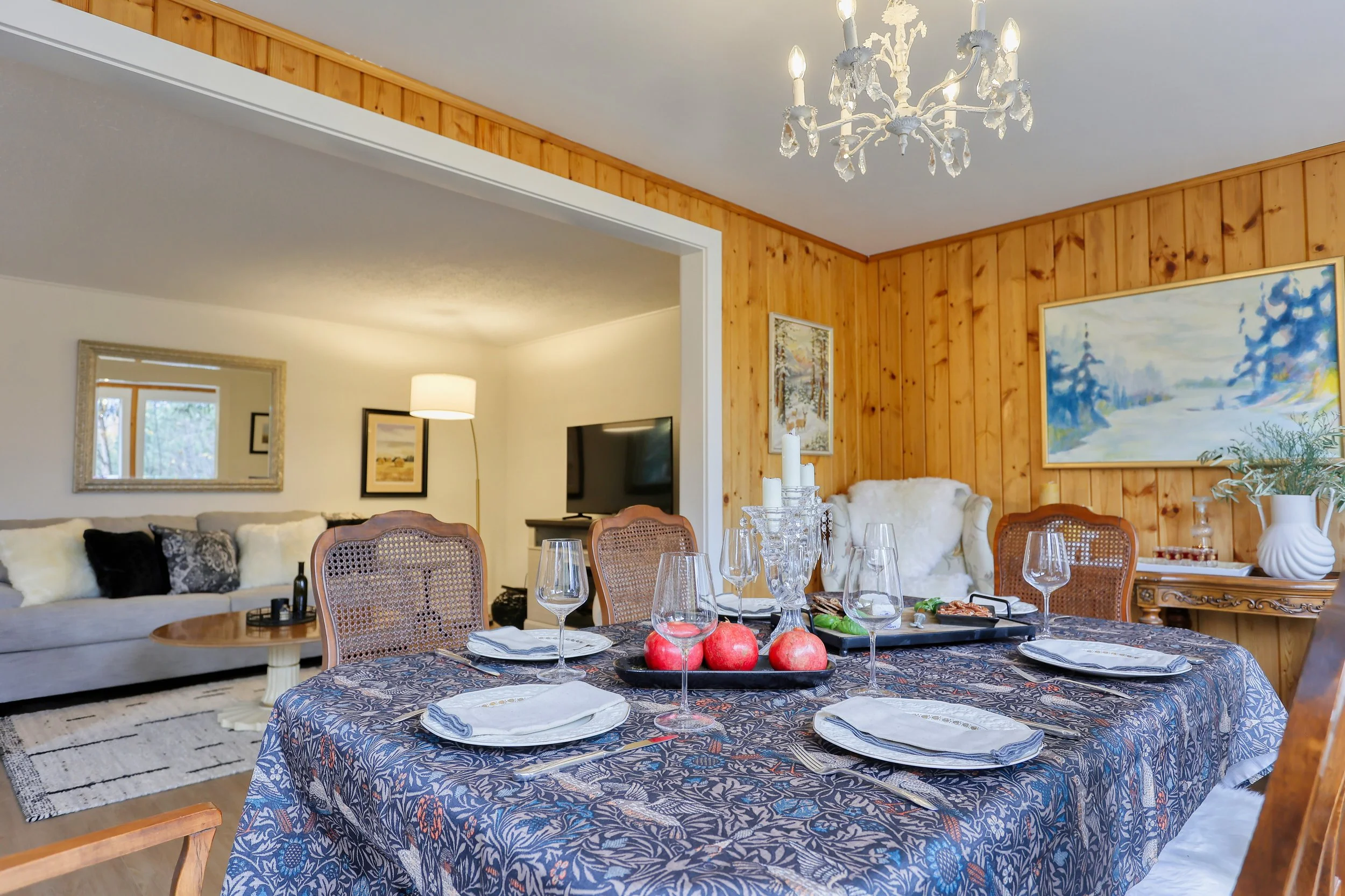 A cozy dining and living area with a wooden accent wall. The dining table is set with a patterned tablecloth, plates, wine glasses, and a centerpiece featuring red apples and candles. A chandelier hangs above the table. The adjacent living room has a