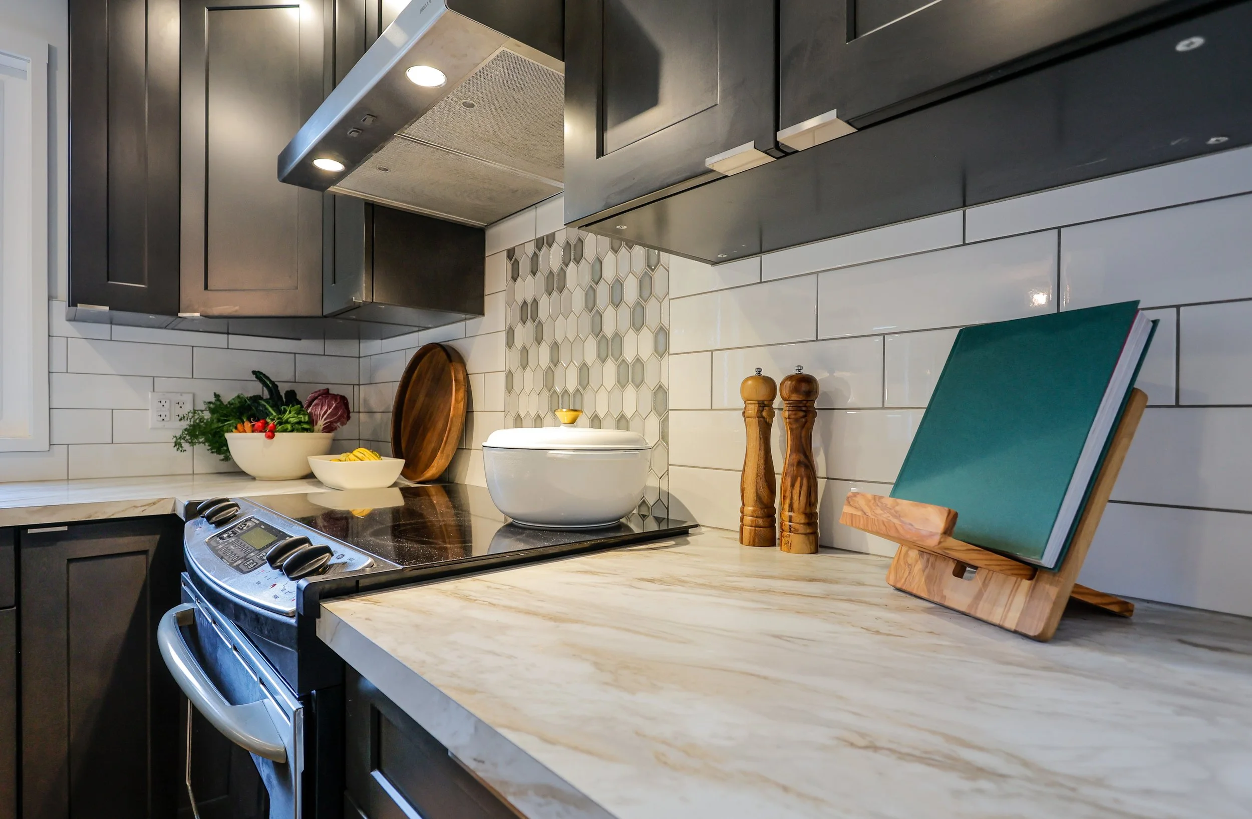 Modern kitchen with dark cabinets, marble countertop, white tile backsplash, and hexagonal accent tiles. Features a stovetop, overhead hood, white pot, pepper mills, cutting board, and open cookbook.