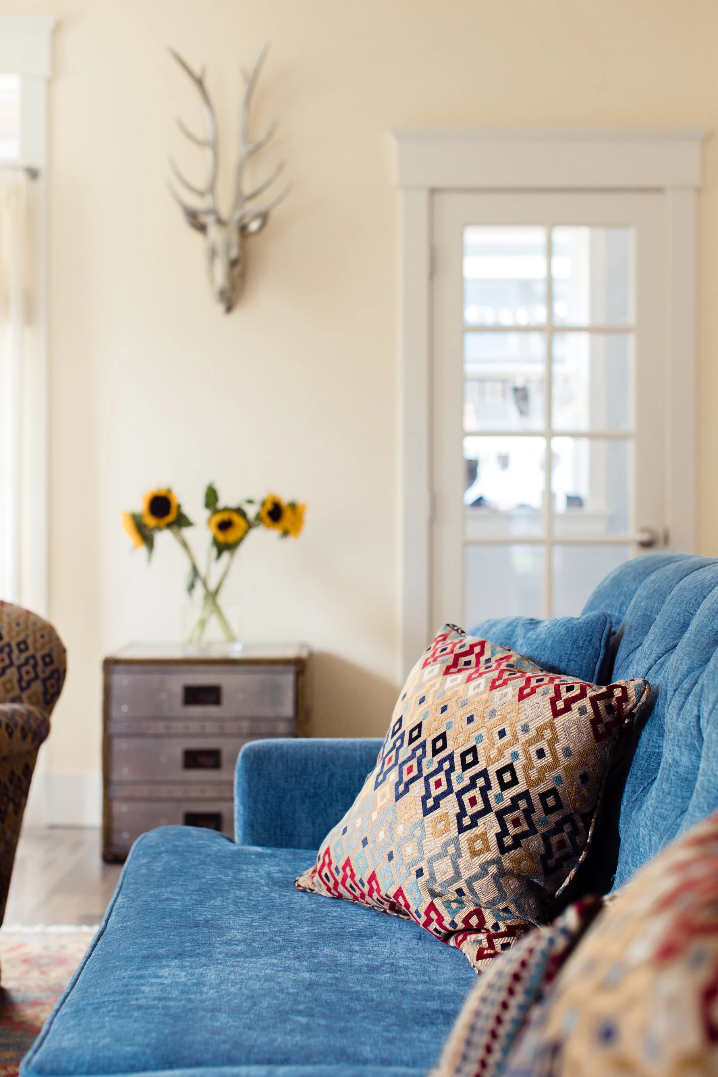 Modern living room with blue sofa, patterned pillows, sunflowers in vase on metal side table, antler wall decor, and glass-paned door.