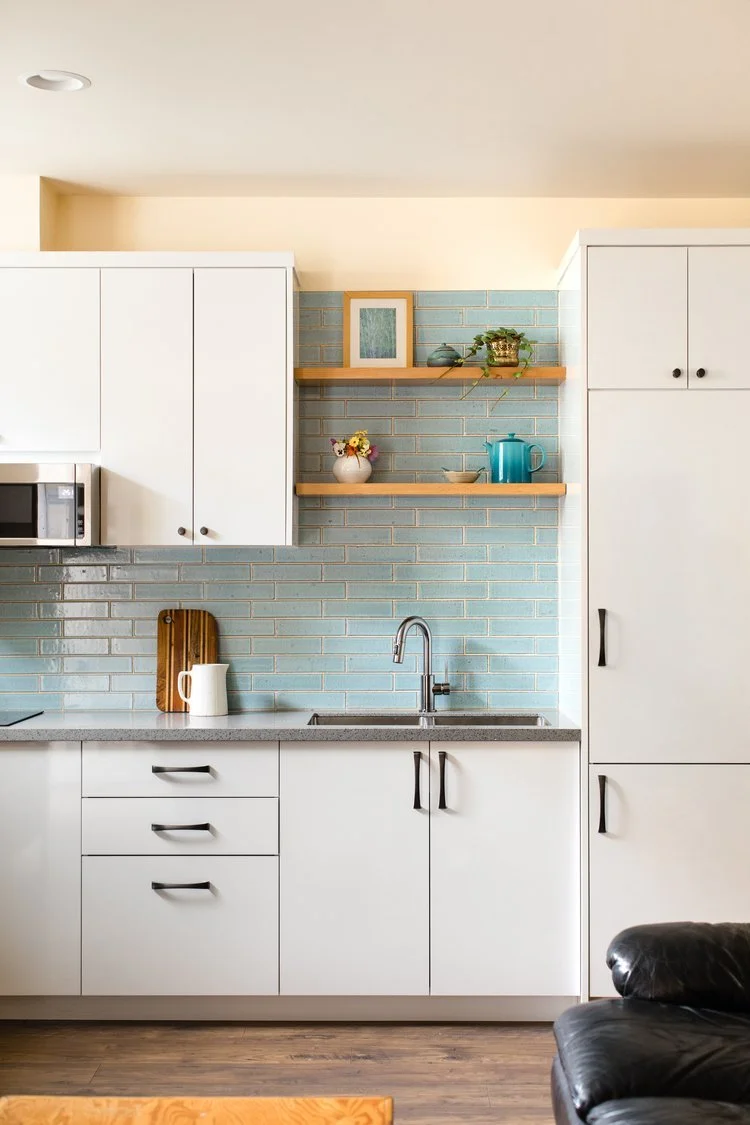 Modern kitchen with white cabinets, blue tile backsplash, and wooden shelves. A sink, faucet, cutting board, teapot, and decor items are visible.