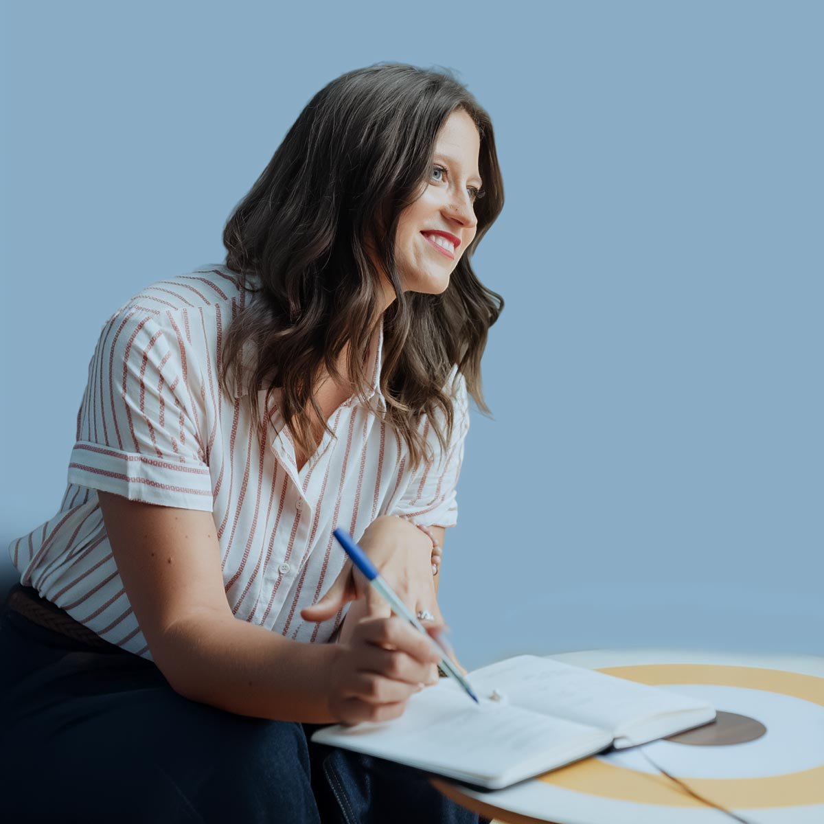 A woman with long brown hair, wearing a striped shirt, smiling and writing in a notebook, sitting at a round table with a blue background.