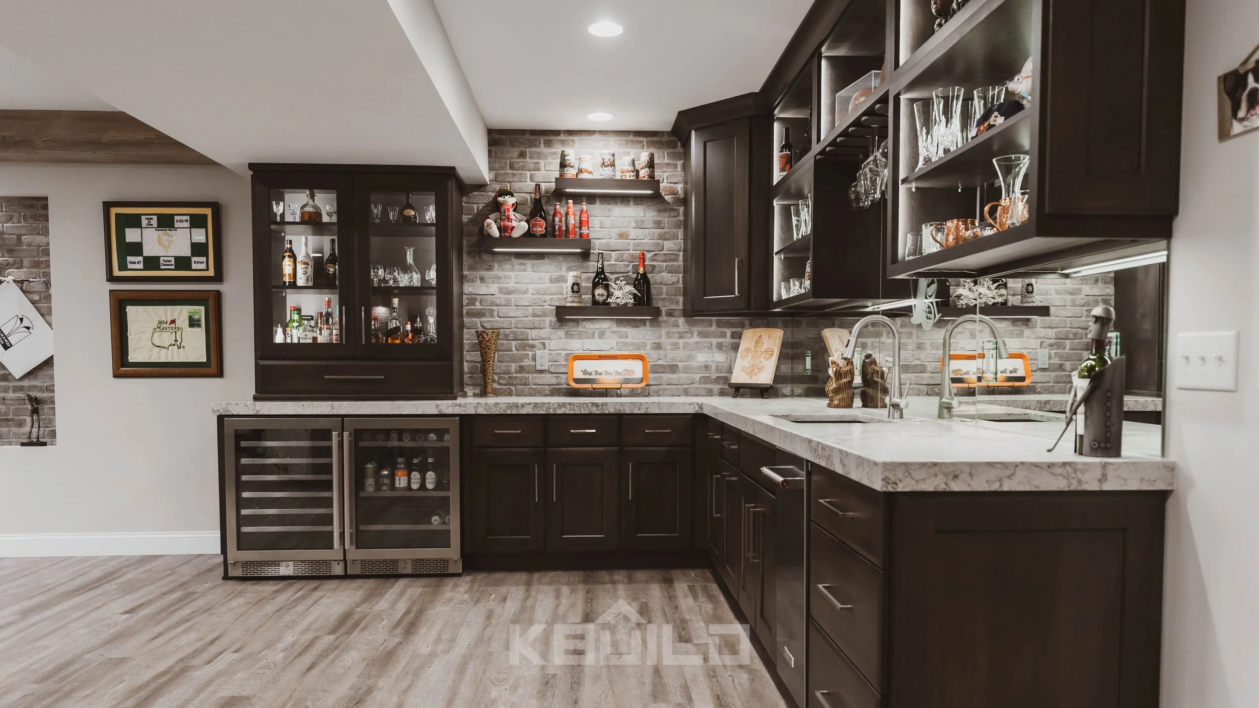 A corner basement bar with dark wood cabinets, light stone countertops, a mini fridge and sink, mirrored and brick backsplash and floating shelves