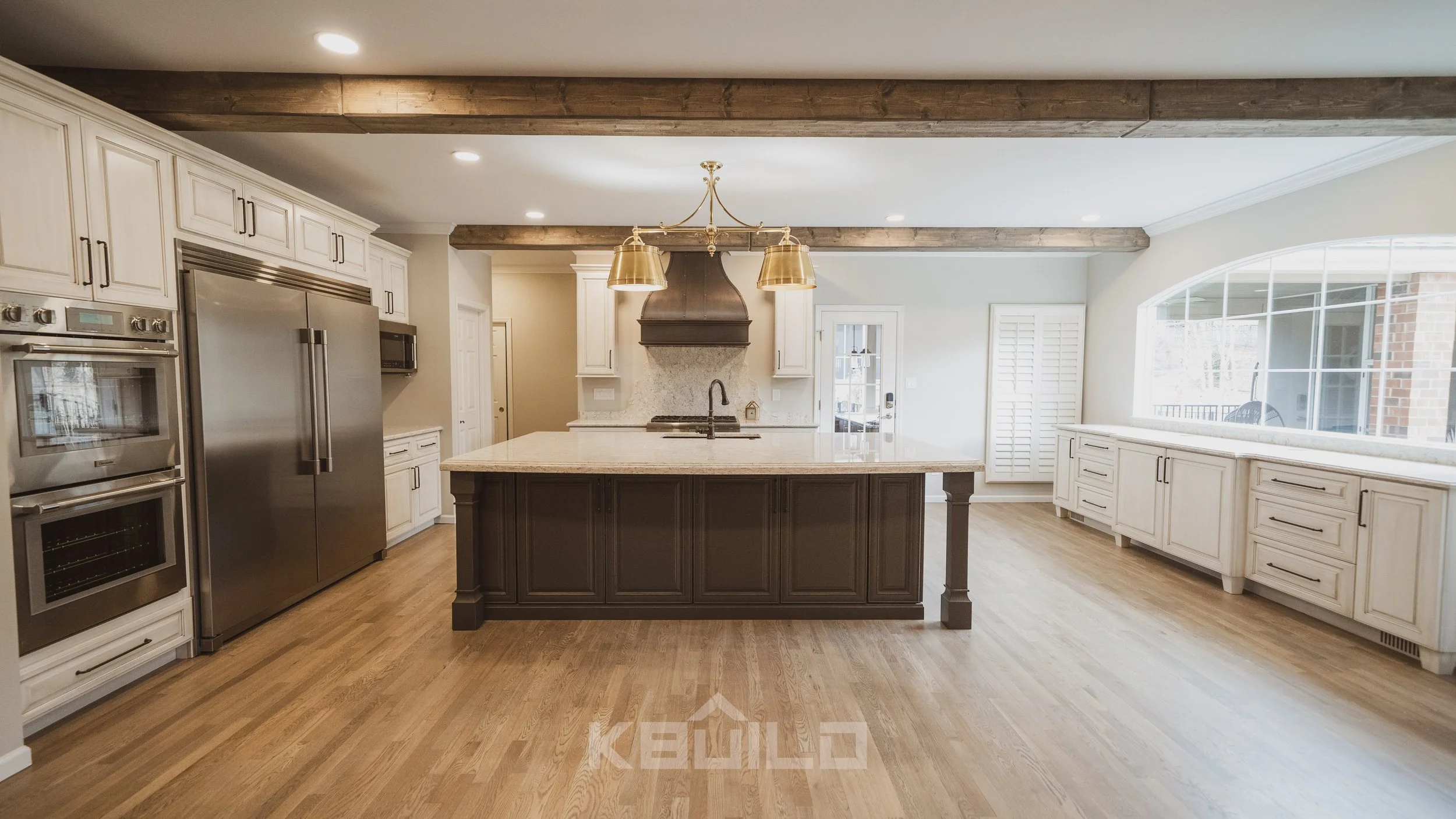 Kitchen remodel with wood flooring, dark kitchen island with white countertops, exposed ceiling beams, and gold hanging light fixtures