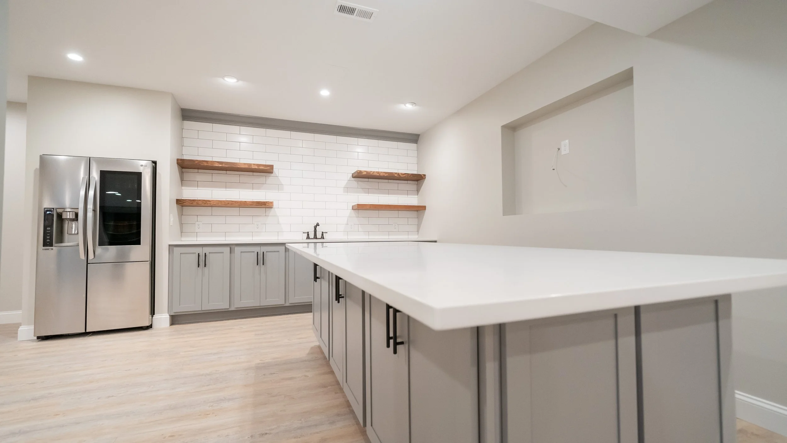 Basement full kitchen with gray cabinets and white stone countertops, subway tile, floating shelves, full fridge and microwave