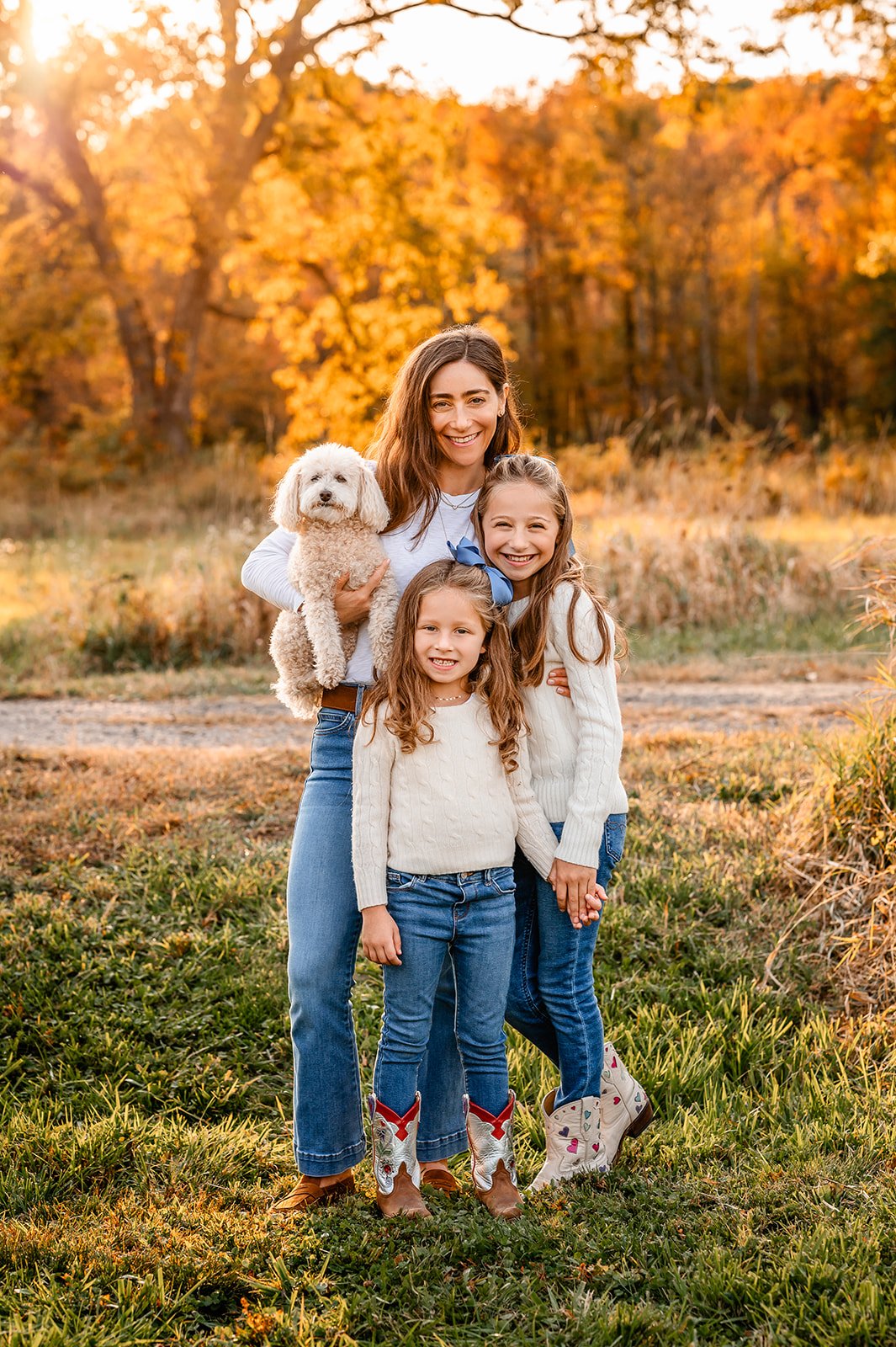 A mom stands with her two daughters and holds a small white dog while they pose for family photos in the fall