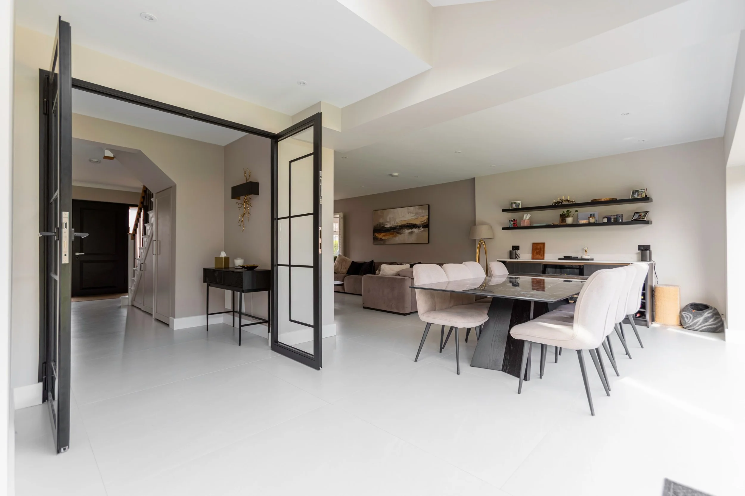 Black steel framed double doors opening into a modern dining and living space with large white floor tiles and contemporary interiors.