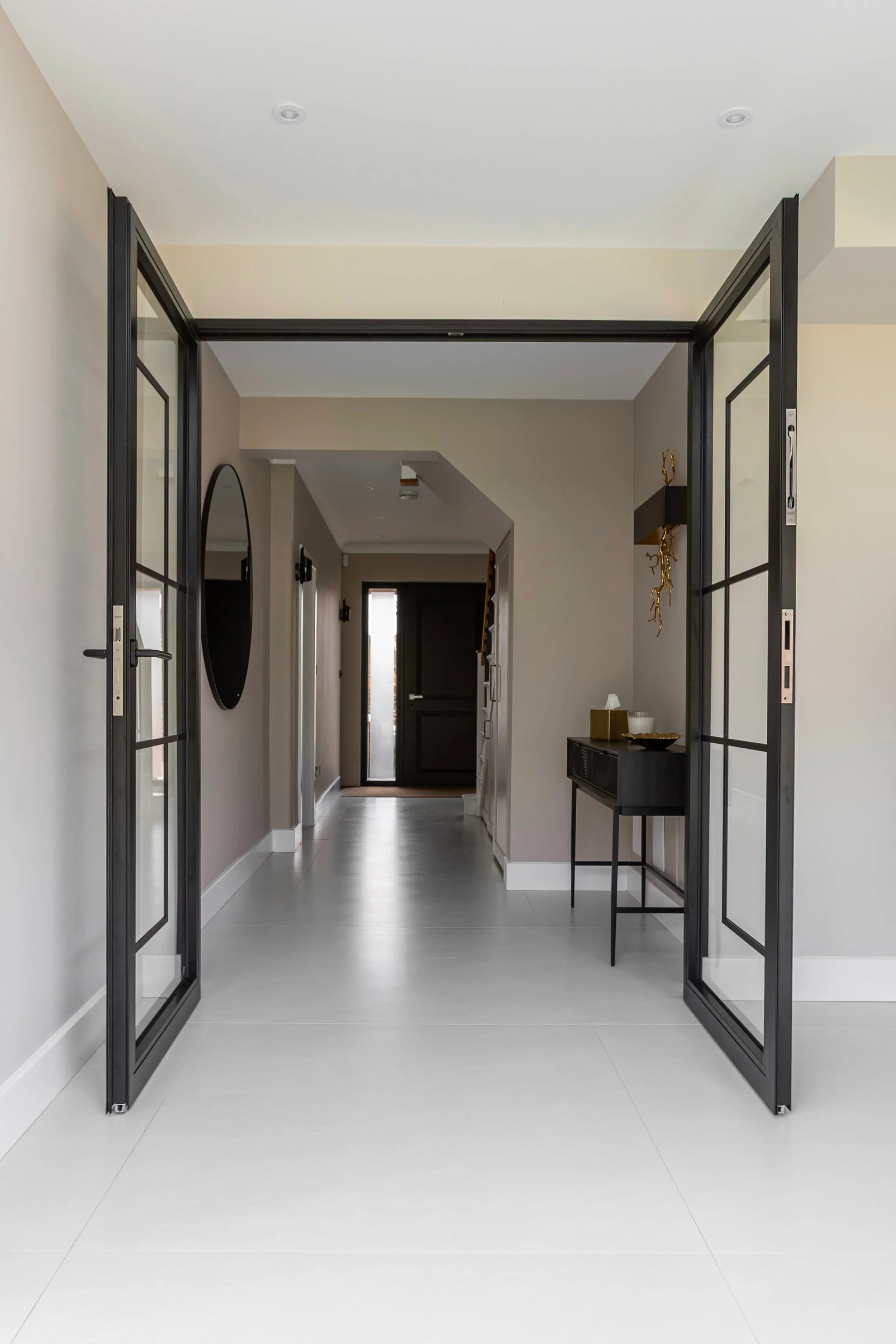 Black steel framed double doors viewed from a hallway, opening into a contemporary open-plan home with light tiled flooring.