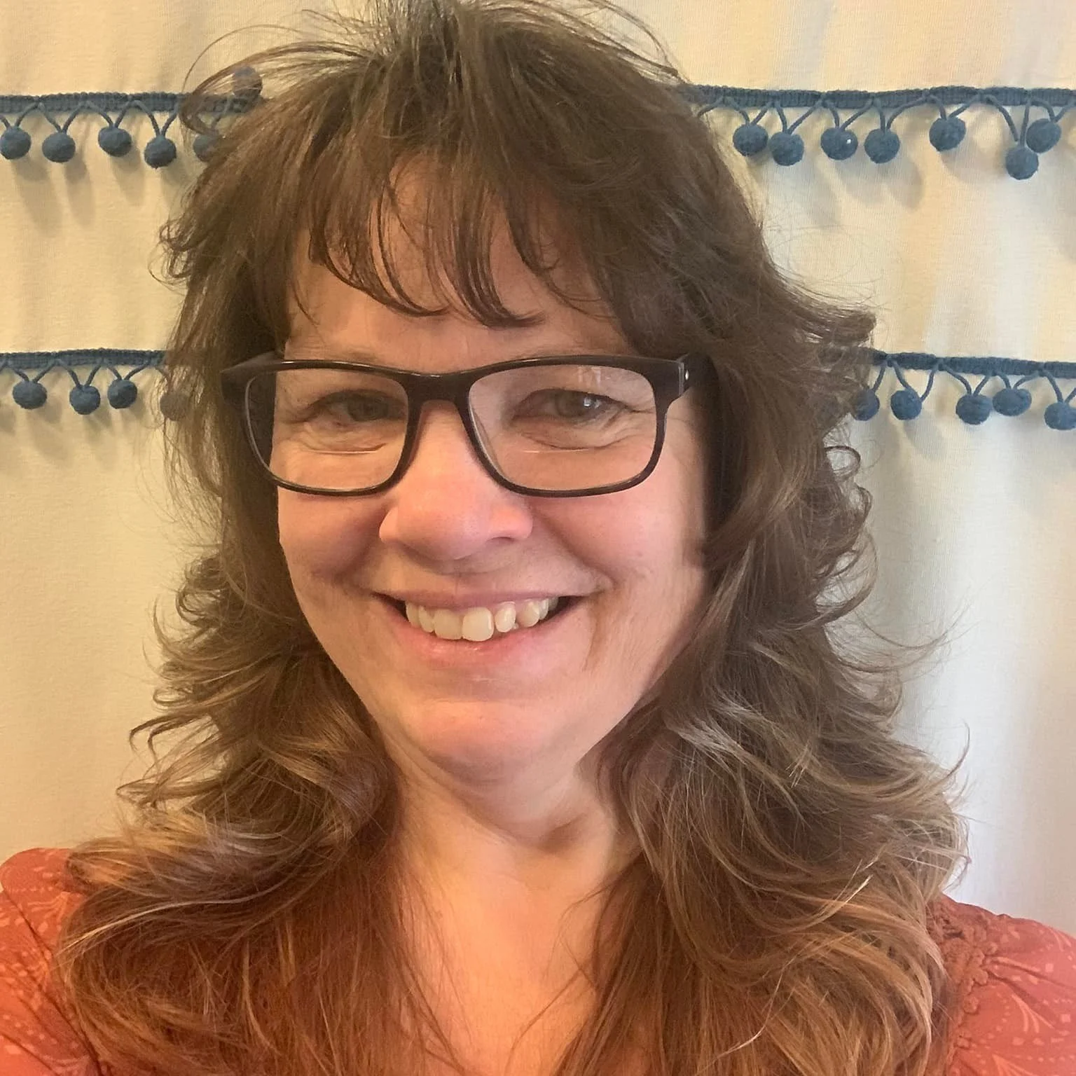 A woman with brown, curly hair and glasses smiling at the camera in front of a beige curtain with blue pom-pom trim.