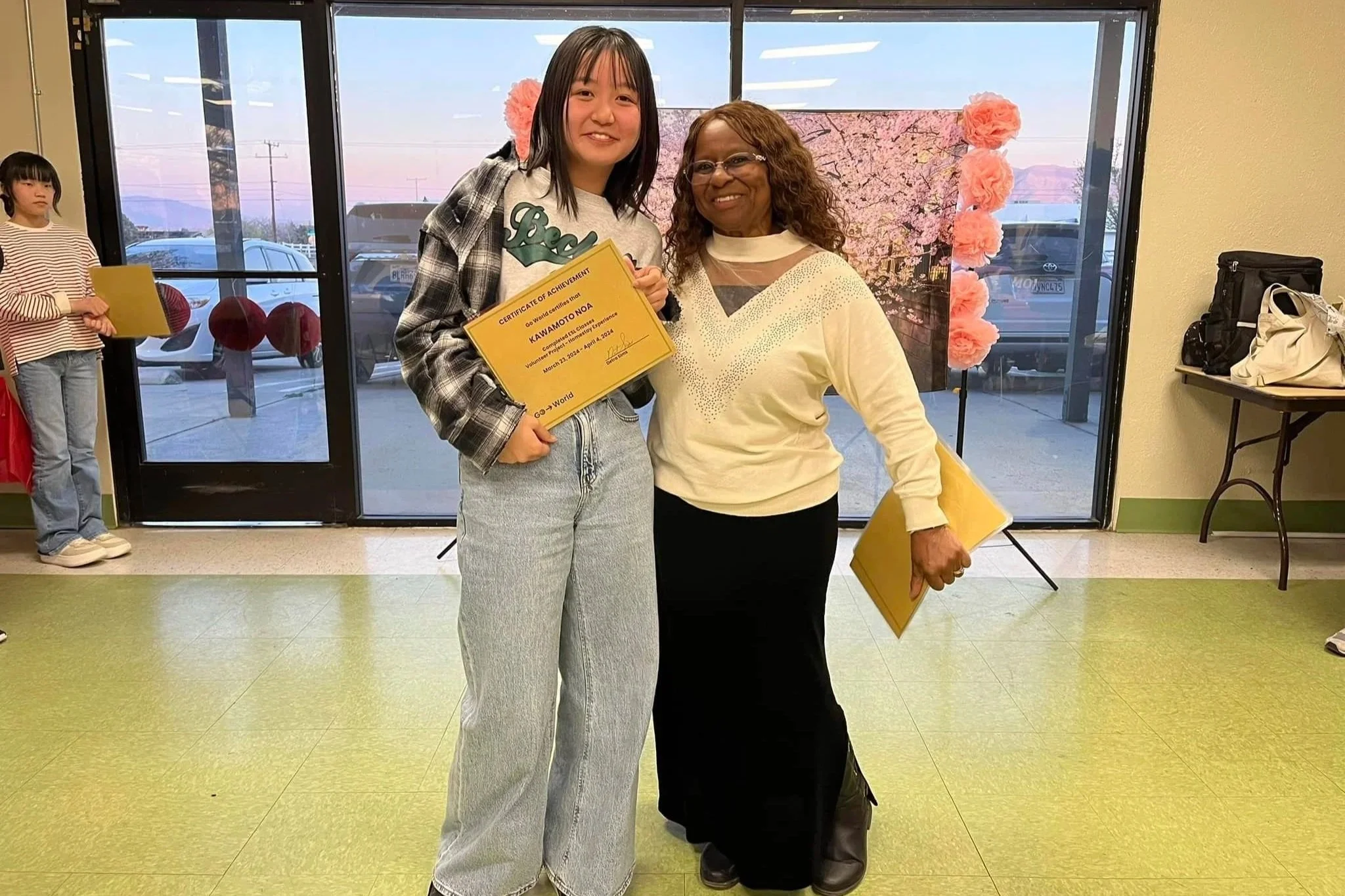 Two women standing indoors, smiling, holding certificates. The woman on the left is younger with dark hair, wearing a plaid jacket and light jeans. The woman on the right is older with curly hair, wearing glasses, a cream sweater, and black pants. Behind them is a backdrop with pink flowers and a window showing a parking lot outside.
