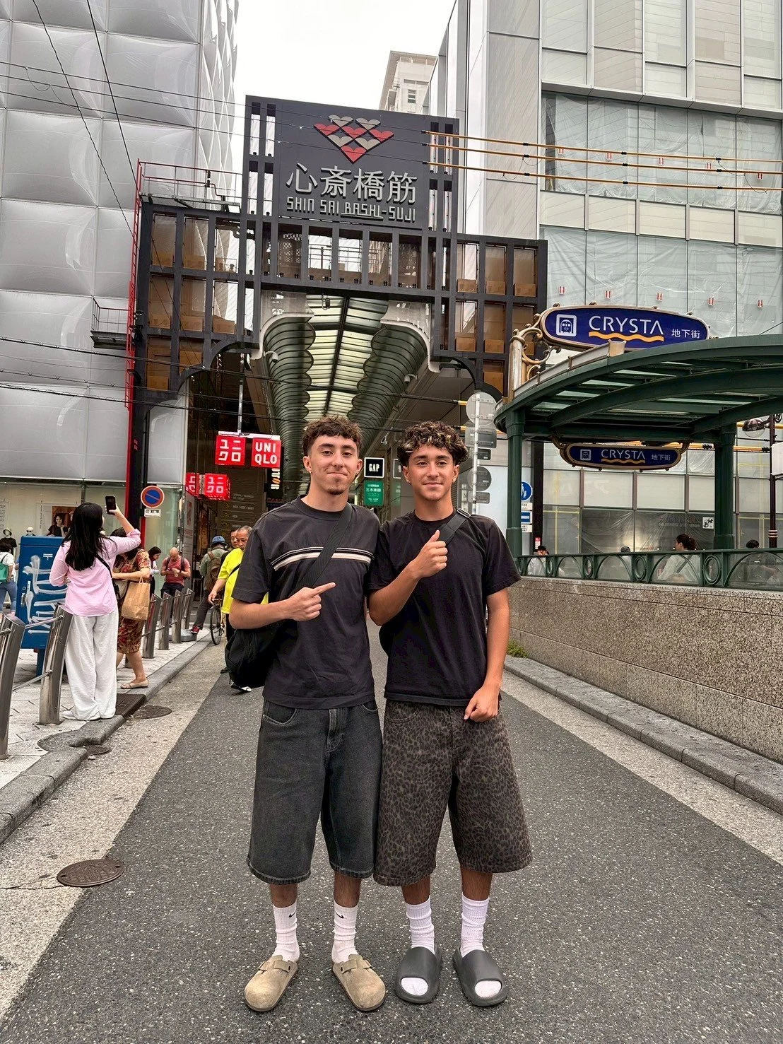 Two young men stand on a busy city street in front of a shopping entrance, pointing at each other. They are dressed casually in black t-shirts, shorts, and socks with slides. Behind them is a street sign, buildings, and other pedestrians.