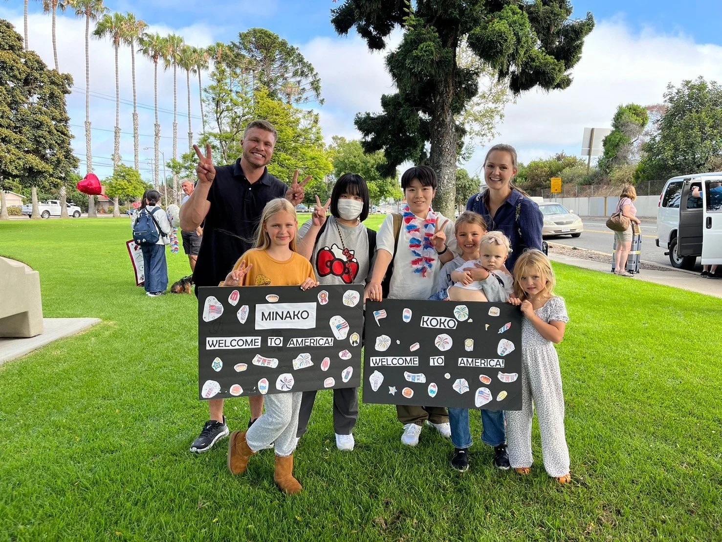 A group of children and two adults standing on a grassy area holding welcome signs that read 'Minako Koko Welcome to America!' in a park, with trees, cars, and other people in the background.
