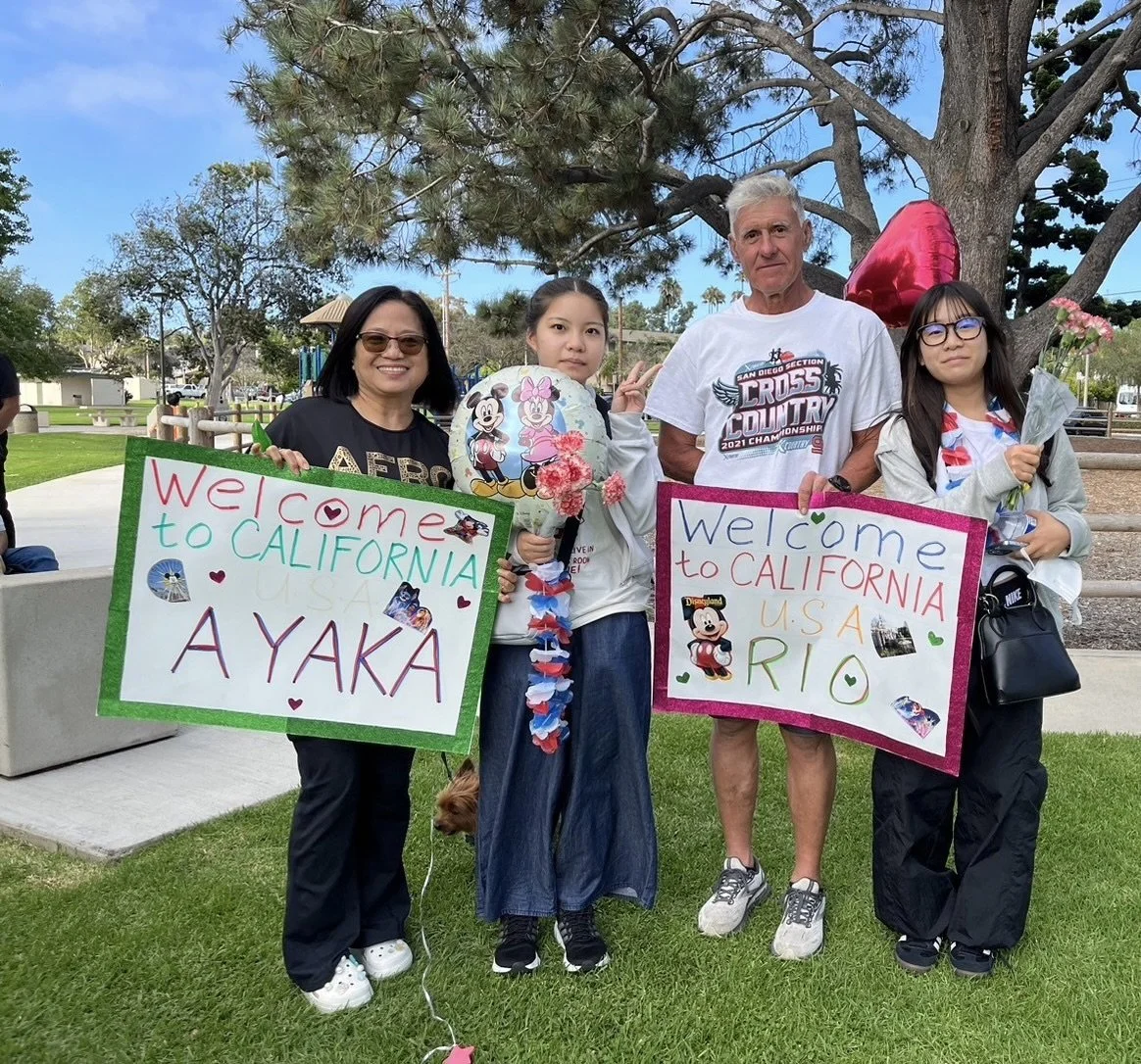 Four people standing outdoors holding welcome signs for California, with two young girls holding balloons and flowers. The signs say "Welcome to California Ayaka" and "Welcome to California Rio," decorated with stickers and hearts. They are under trees in a park-like area.