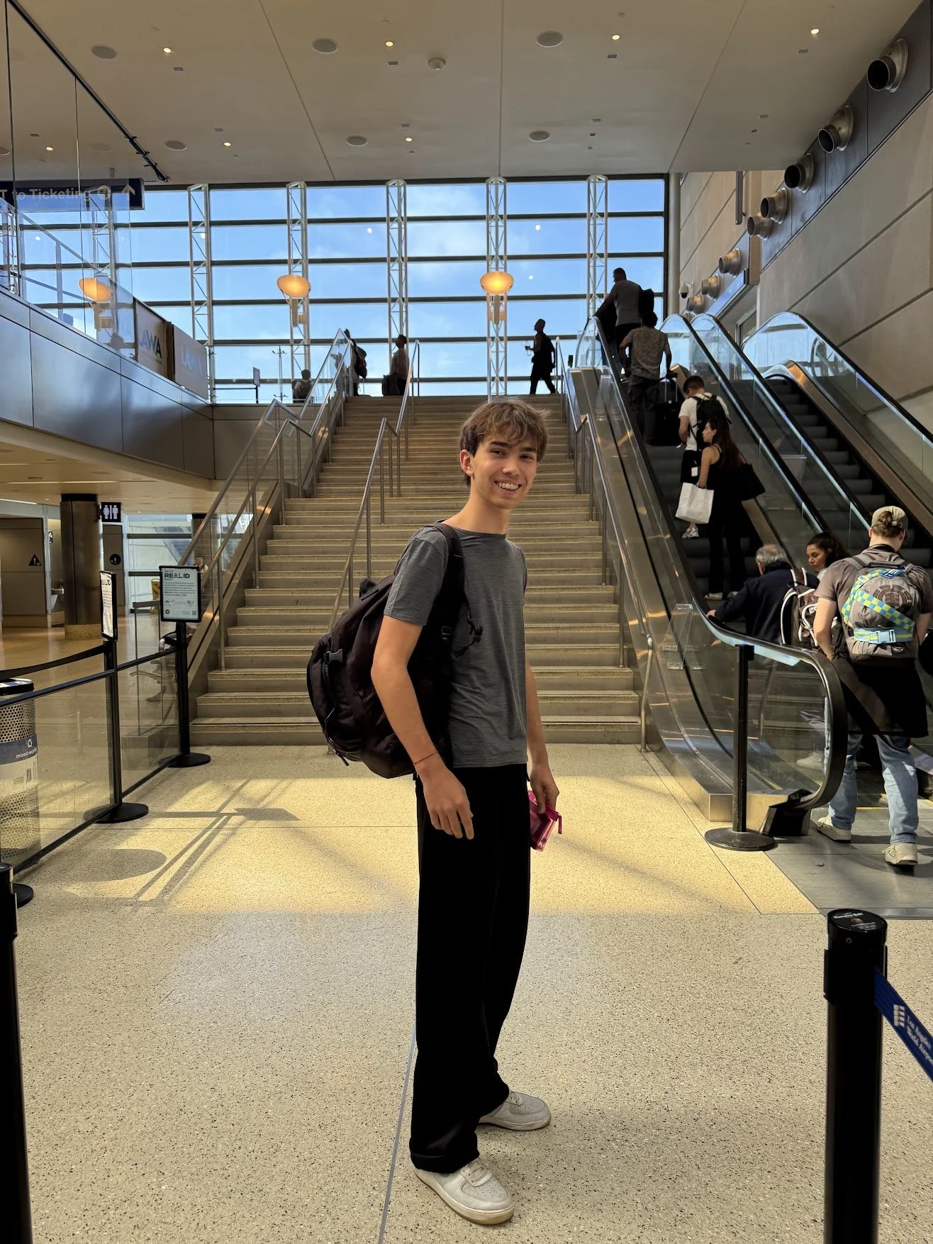 A smiling young man with a backpack standing inside an airport terminal near escalators, with travelers walking and sitting in the background.
