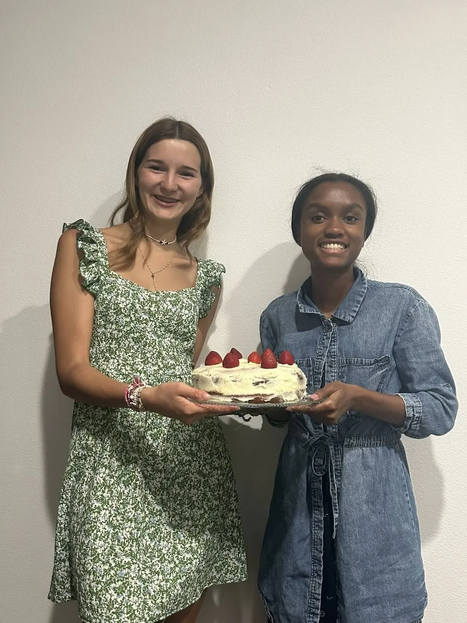 Two young women standing against a white wall, holding a birthday cake with strawberries on top and smiling.