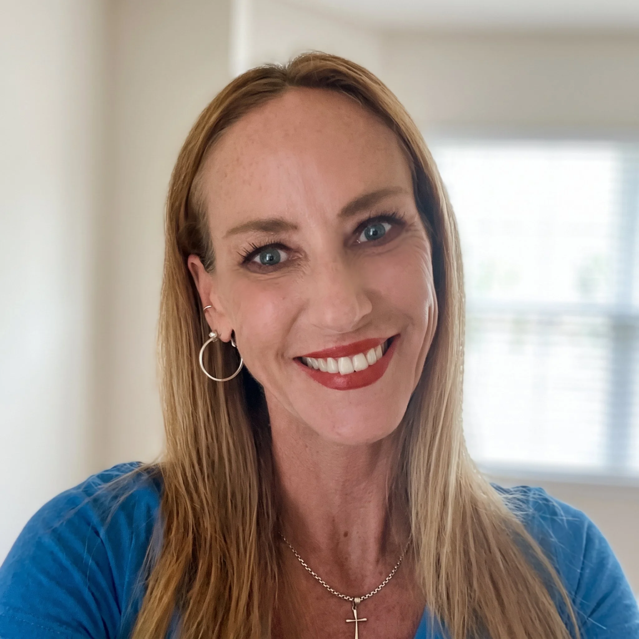 Jodie Shrode, a Licensed Mental Health Counselor at Dandelion Therapy Group in Fort Myers, smiles warmly in a professional headshot, wearing a blue top and silver cross necklace against a softly blurred, neutral indoor background.