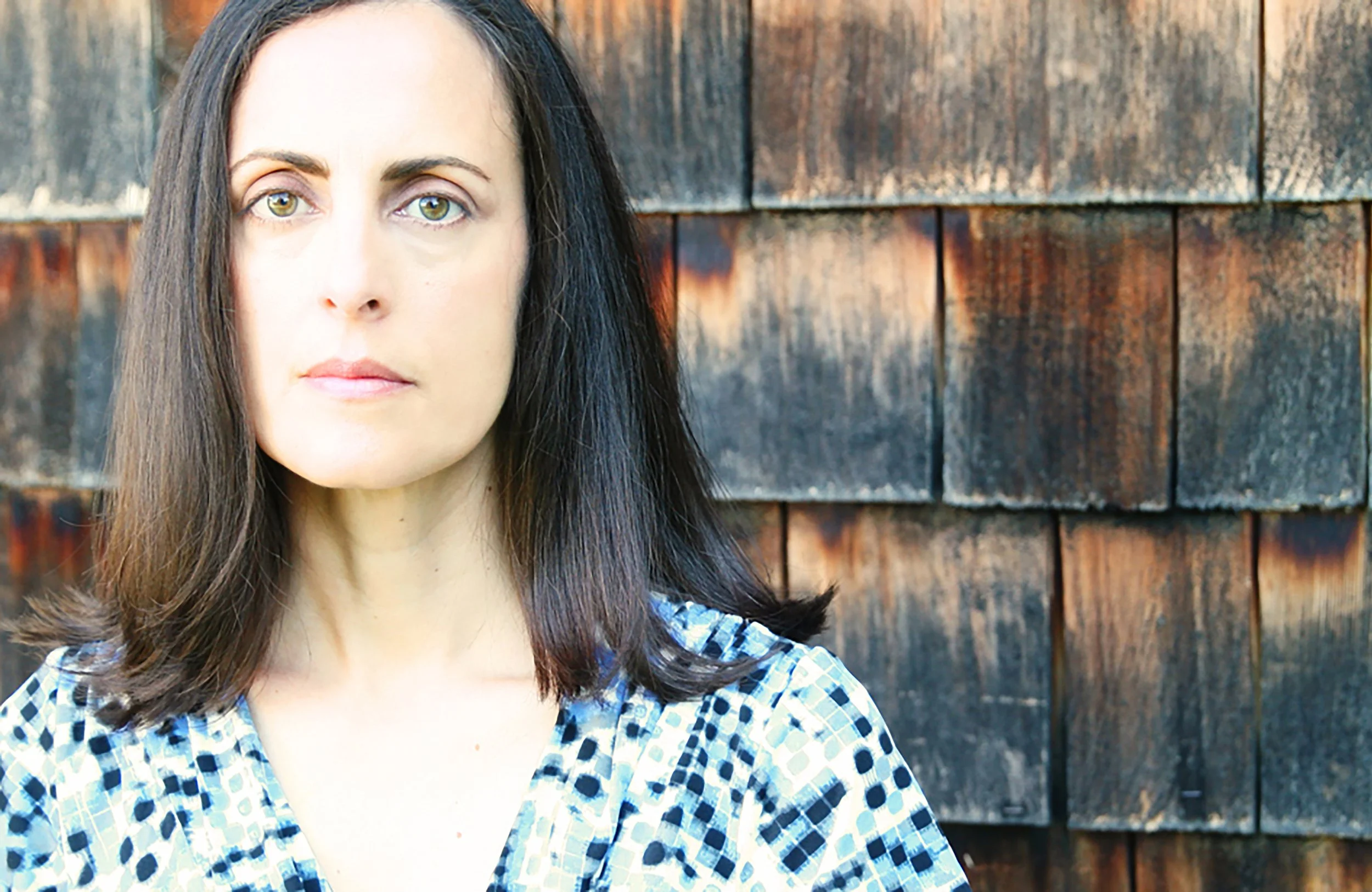 A woman with long, dark hair stands in front of a wooden shingle wall, wearing a patterned blue and white blouse.