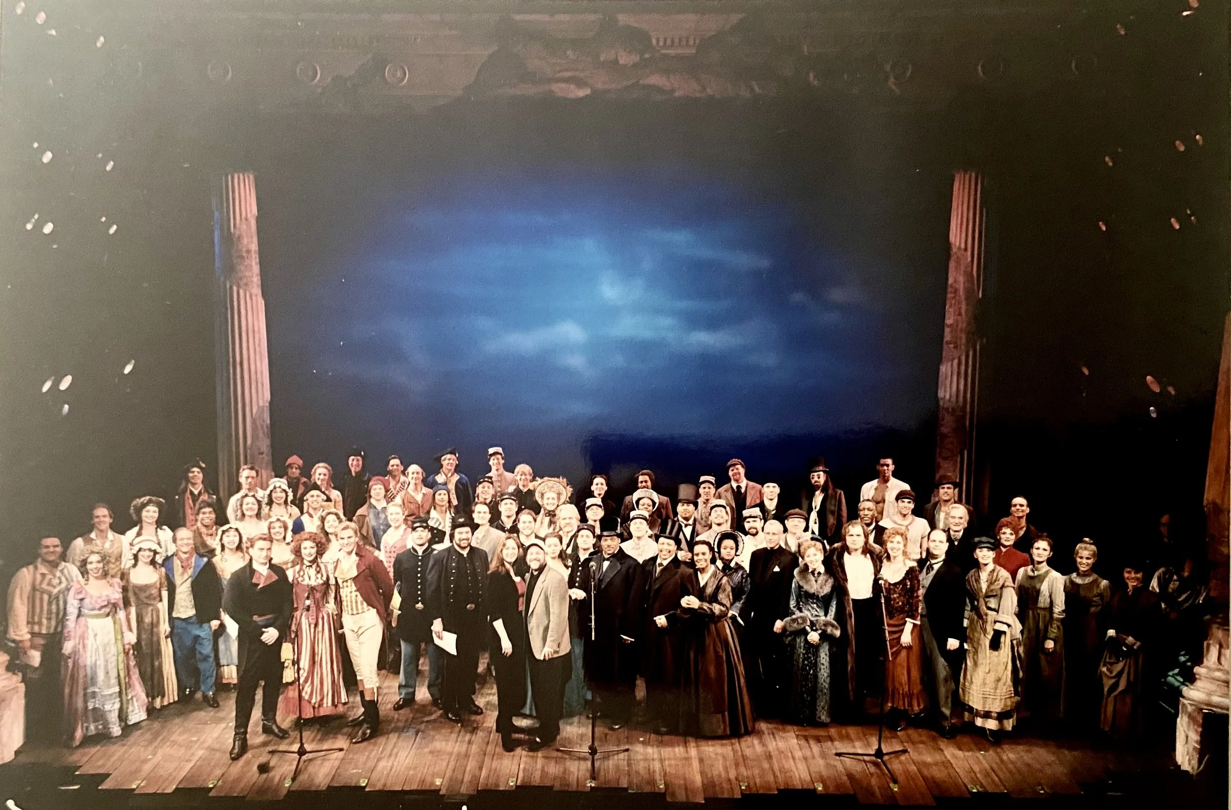 A large group of people dressed in historical costumes on a stage with a dark, cloudy sky backdrop and two tall columns on either side.