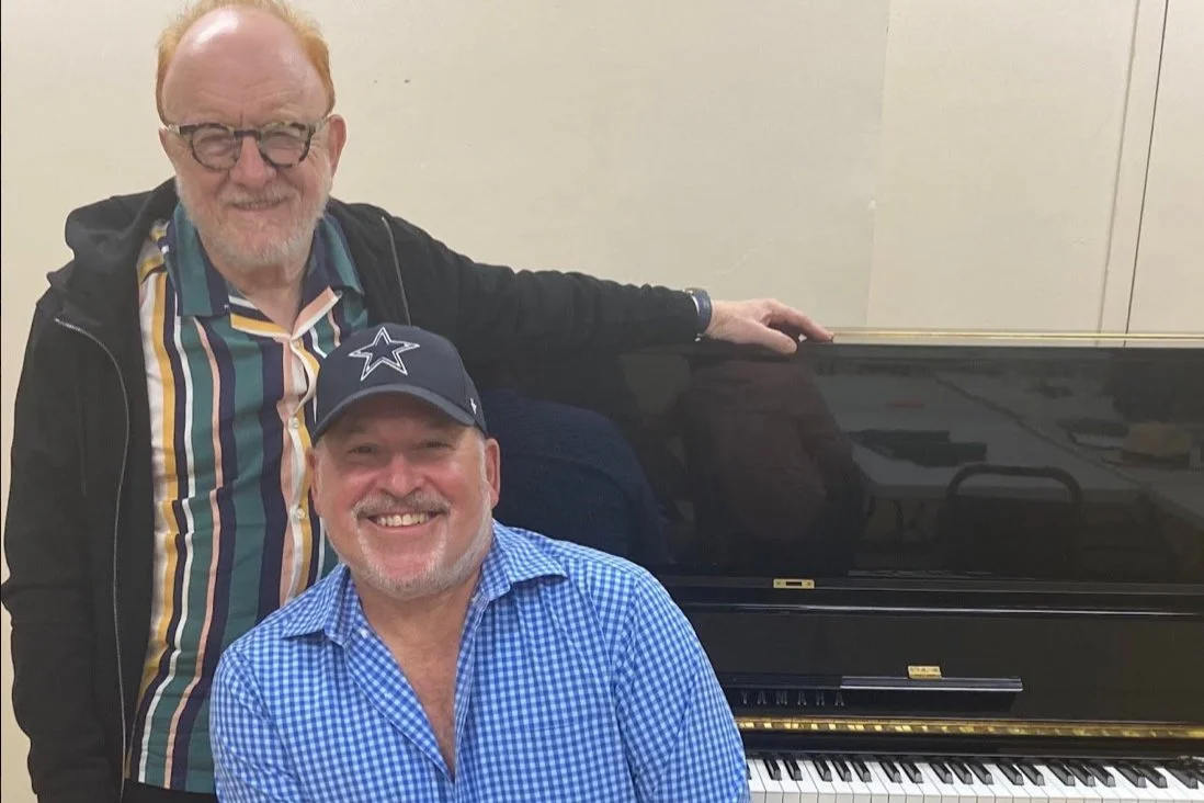 Two smiling men, one standing and one sitting, in front of a black Yamaha piano. The standing man has red hair and wears glasses, a multicolored striped shirt, and a black jacket. The sitting man wears a Dallas Cowboys cap and a blue checkered shirt.