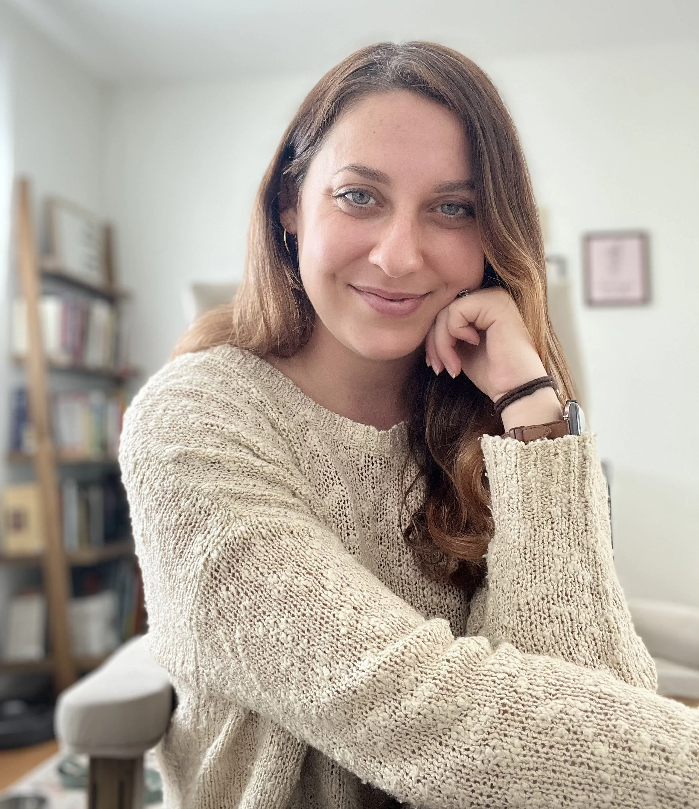 Close-up of a woman with long, wavy brown hair, blue eyes, wearing a beige knitted sweater, earrings, and a watch, sitting indoors with a bookshelf and framed picture in the background.