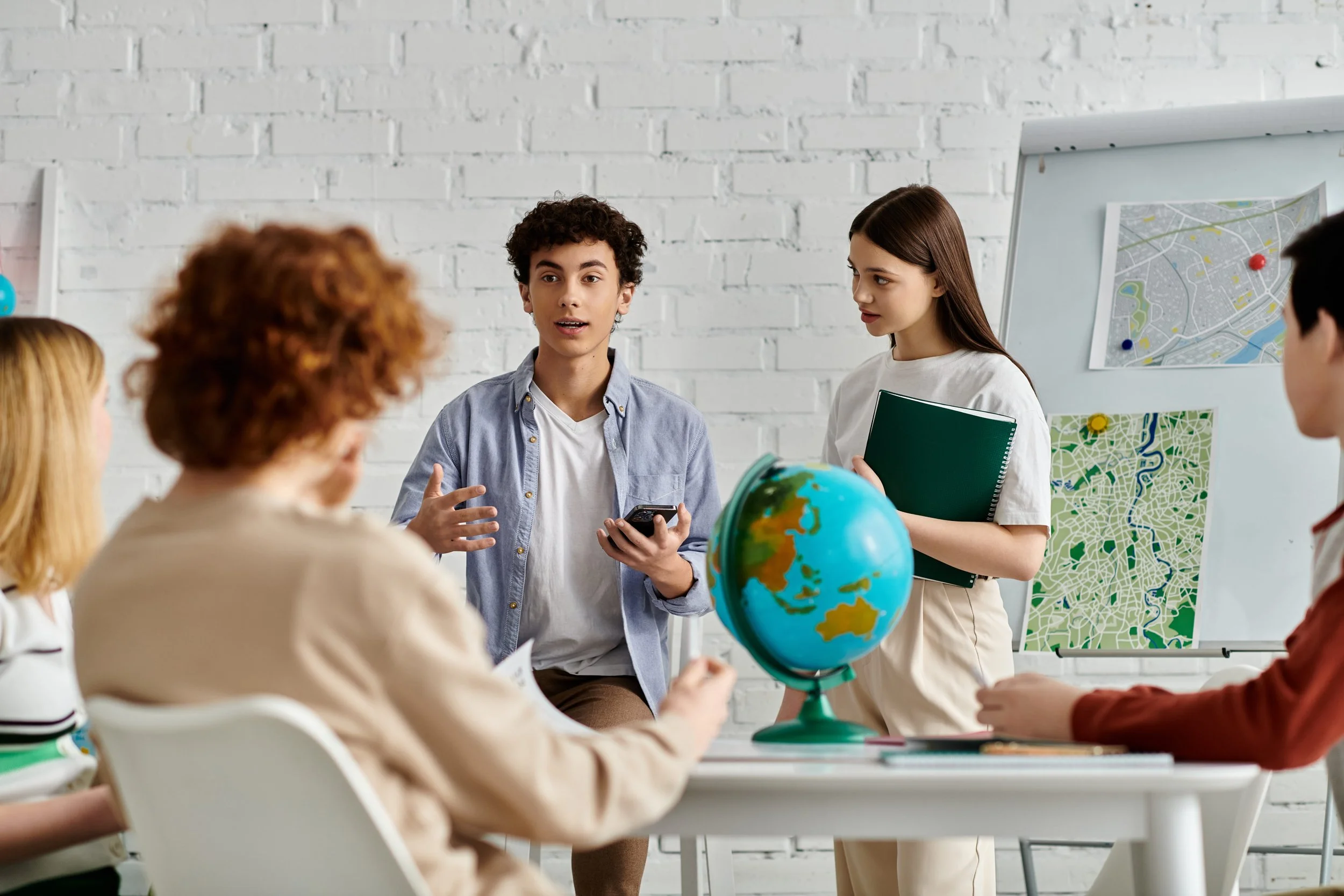Young man in a light speaking in front of model un classroom with a globe and maps of the city on a whiteboard.