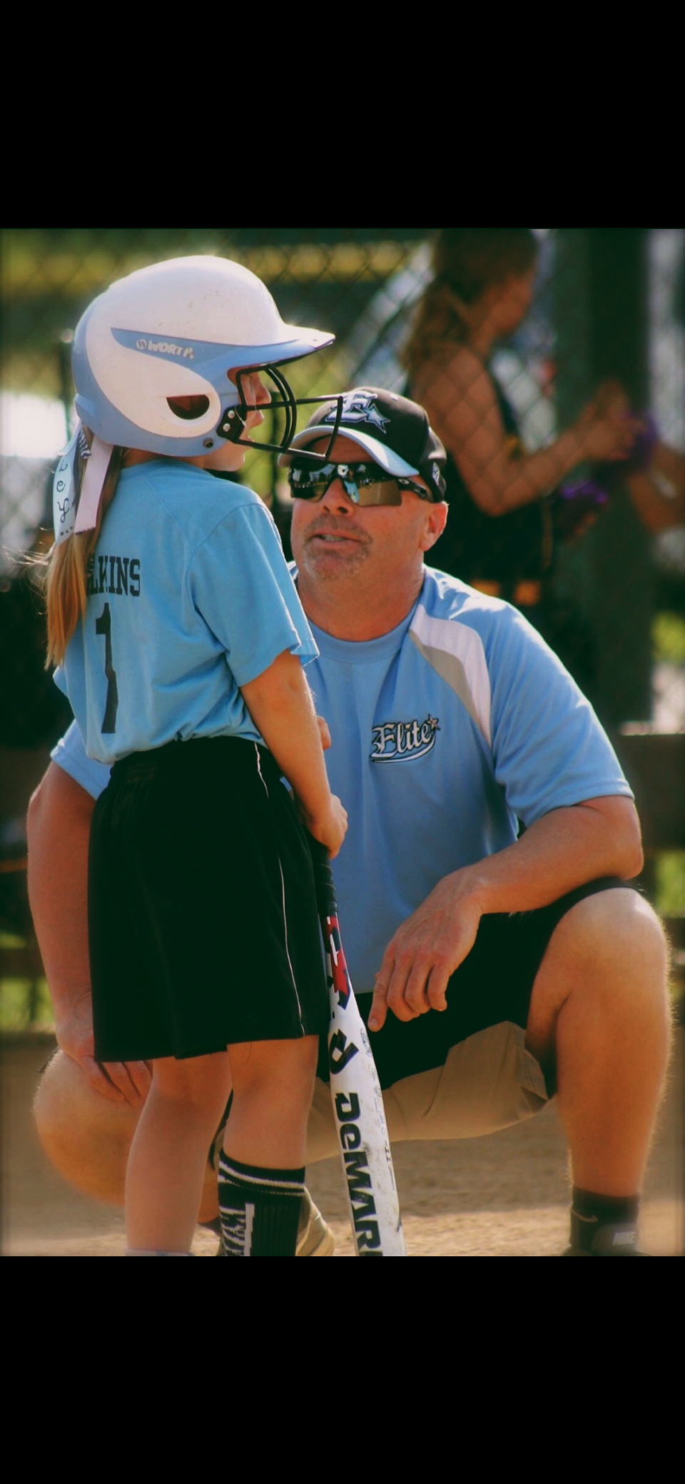 A young girl wearing a baseball helmet and uniform standing on a baseball field, talking to a coach Kelly who is kneeling down.