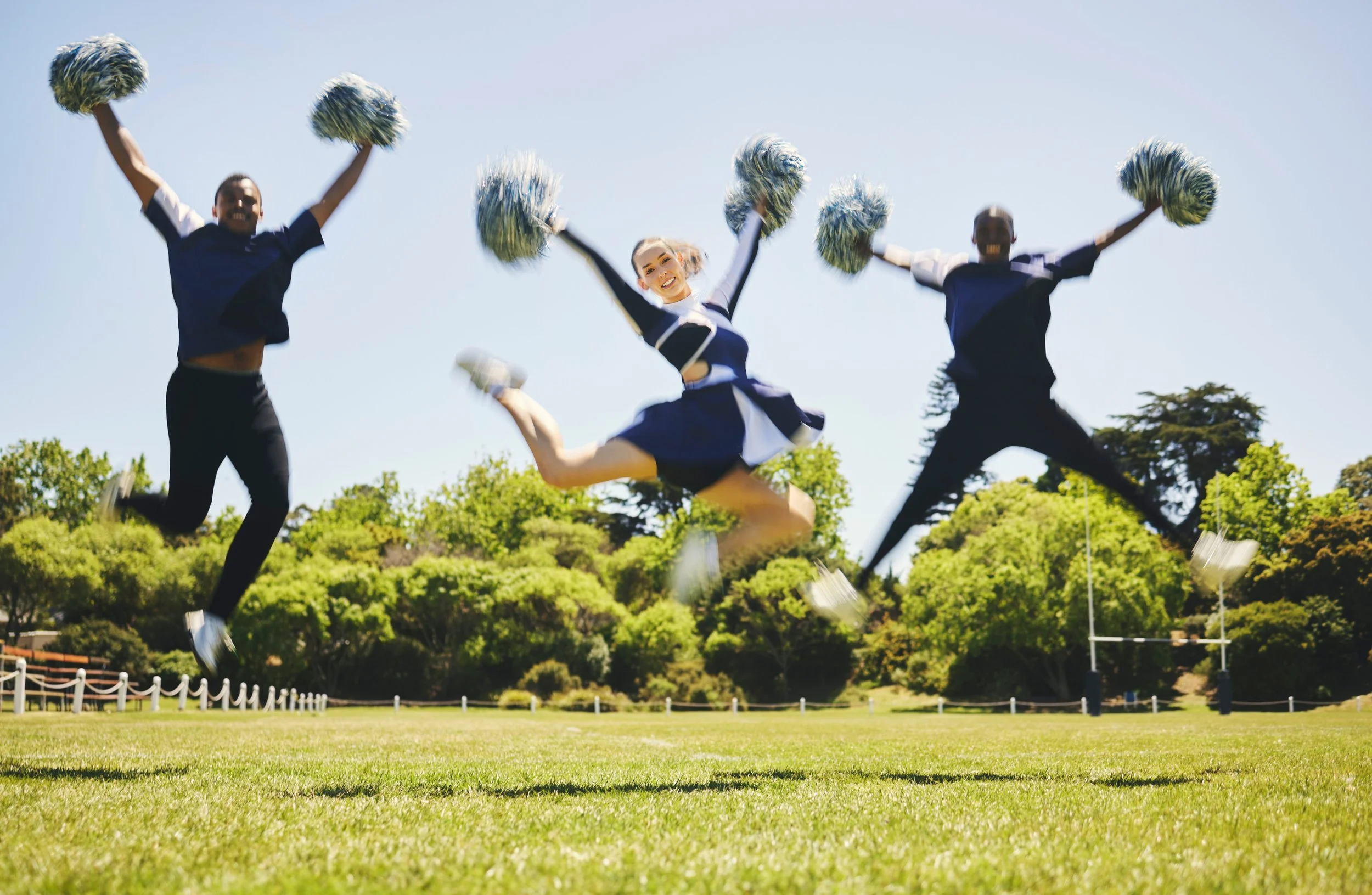 Three cheerleaders jumping in the air outdoors on a sunny day, with the middle one dressed in a cheerleading uniform and the others in casual clothing, holding pom-poms.