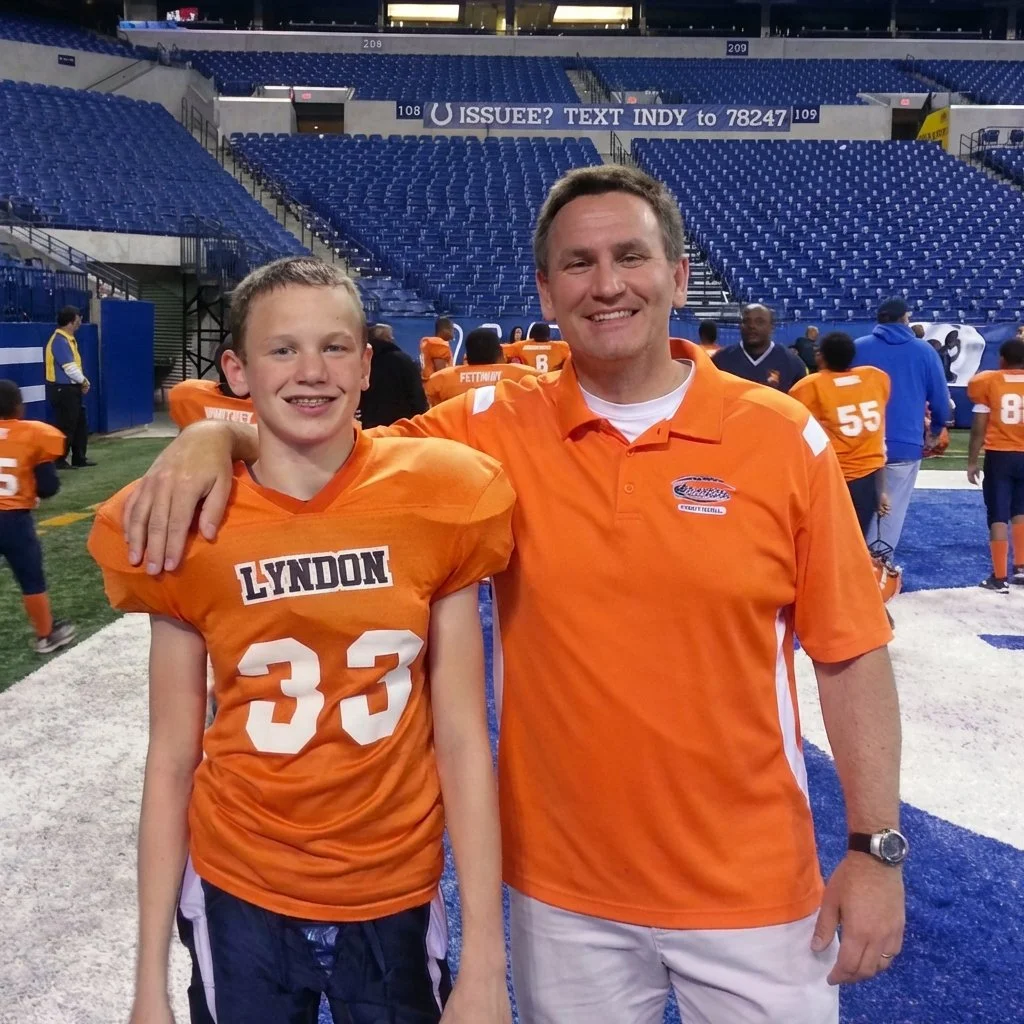 A young boy and a fundraising coach, both wearing orange football jerseys, stand together on a football field at a stadium, smiling at the camera. The boy's jersey reads 'LYNDON 33'.